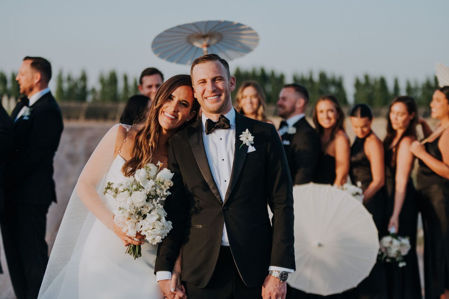 Smiling bride and groom posing with wedding party holding white parasols at sunset.
