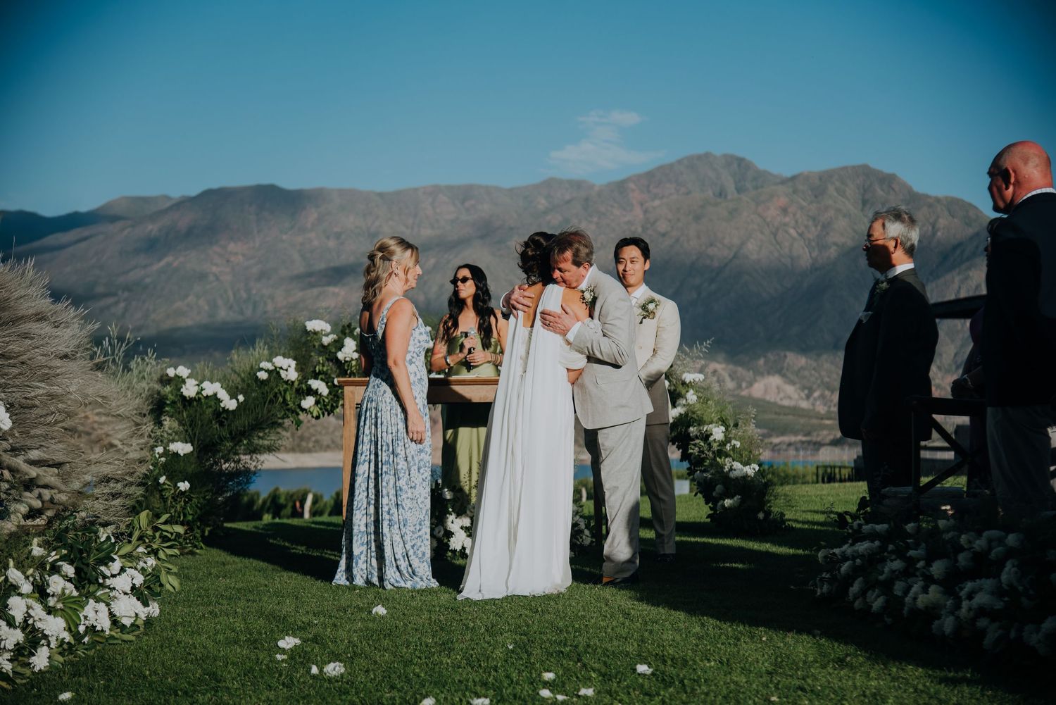 Wedding ceremony outdoors with mountain backdrop, bride and groom exchanging vows surrounded by white floral arrangements.