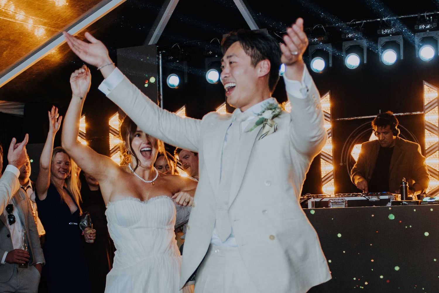 Joyful groom in white suit raises arms while dancing with bride at lively wedding reception.