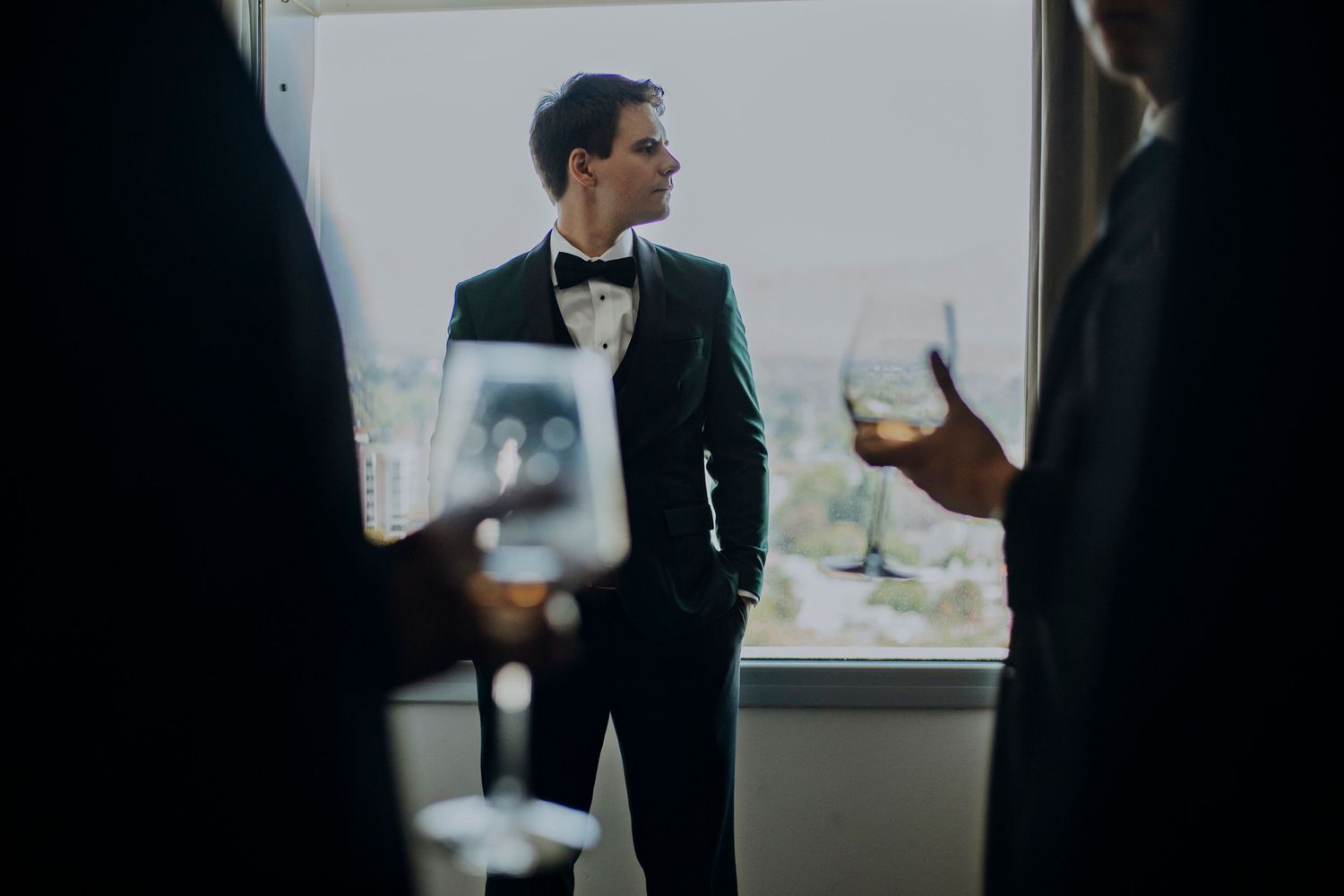 Elegant groom in black tuxedo with bow tie holding whiskey glass near window at upscale wedding reception.