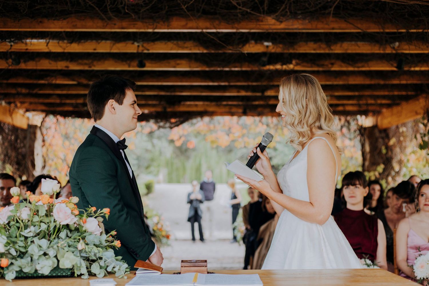 Bride reading vows to groom at outdoor wedding ceremony under wooden pergola.