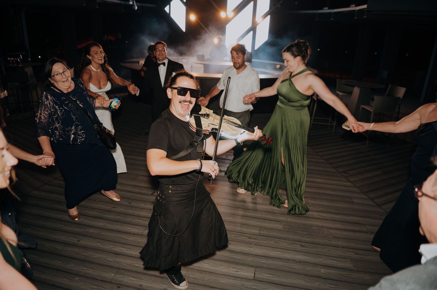 Guests celebrate on the dance floor at a wedding reception, with one man in sunglasses kneeling while others dance around him.