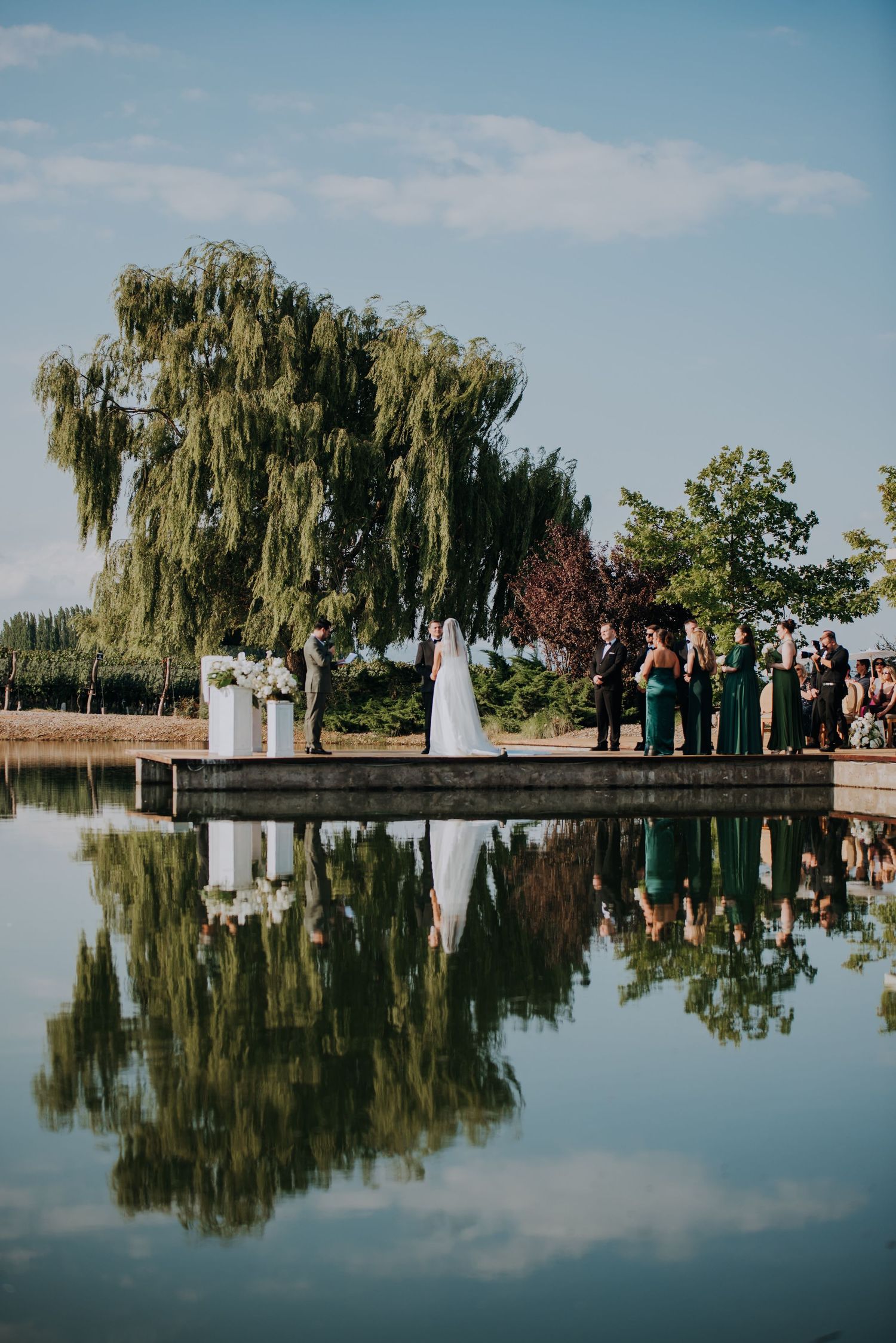Wedding ceremony by reflective pond with willow tree and green bridesmaid dresses.