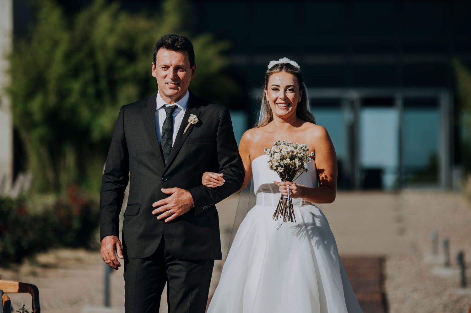 Bride walking down the aisle with her escort, holding a white bouquet at an outdoor wedding.