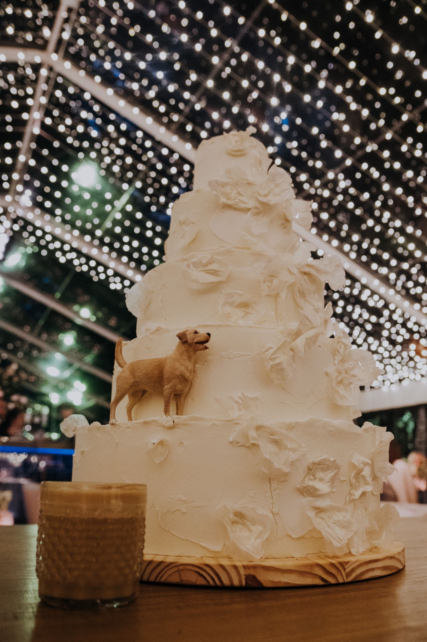 Four-tier white wedding cake with floral details and a golden dog topper under twinkling string lights.