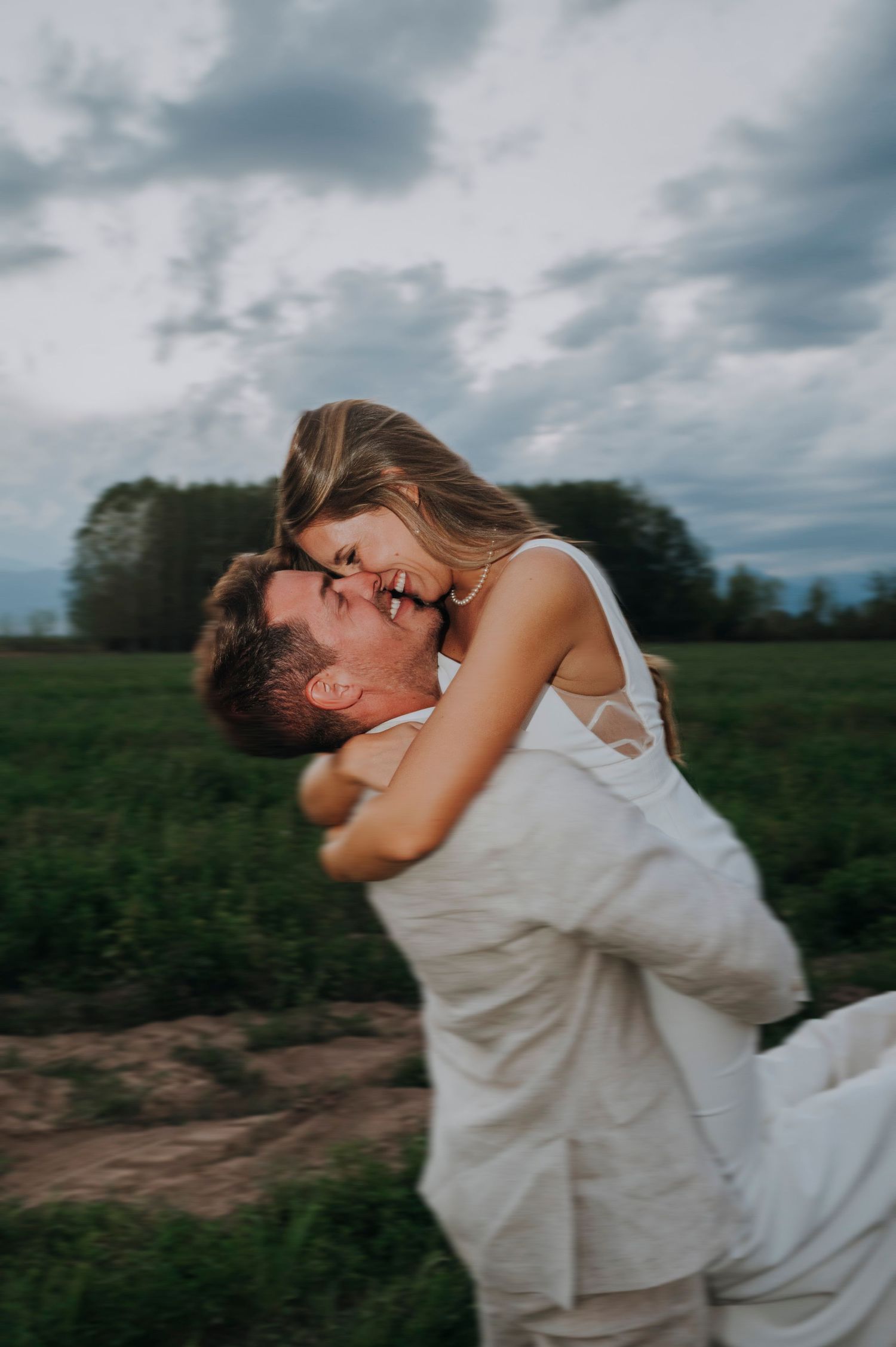 Groom lifts laughing bride in white dress in green field under dramatic cloudy sky. Couple portrait in Mendoza.