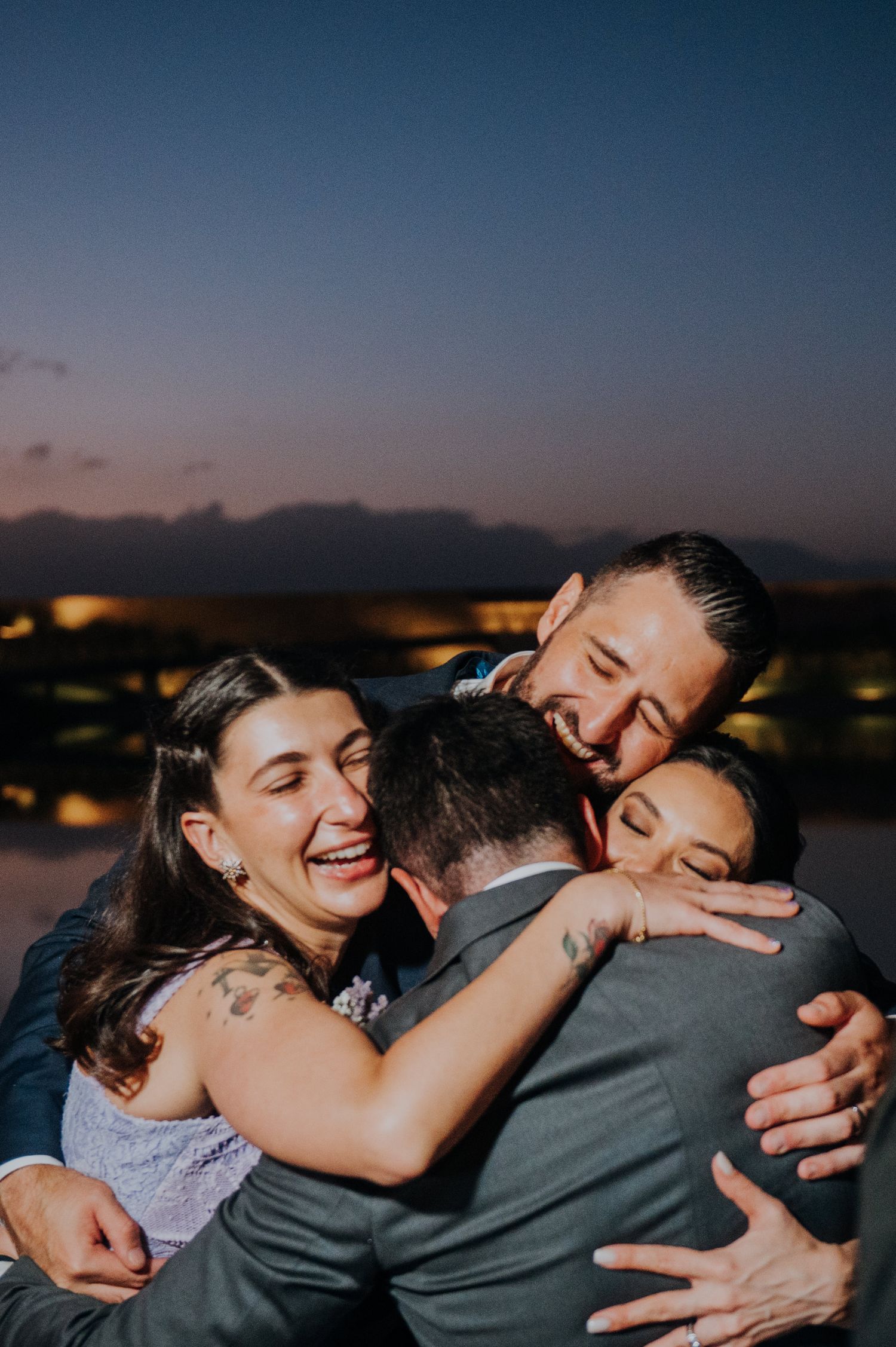 Joyful wedding guests sharing a group hug at twilight with scenic water backdrop, in anaia vineyard mendoza