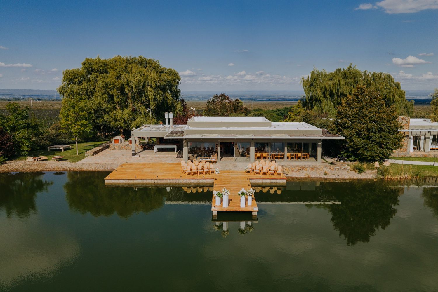 Aerial view of elegant waterfront wedding venue with dock, patio seating, and scenic lake surrounded by trees.