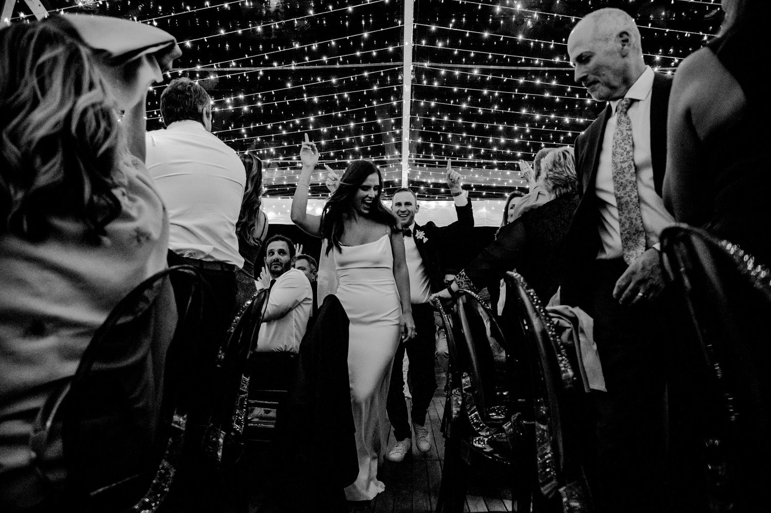 Bride dancing joyfully under string lights surrounded by wedding guests in black and white photo.