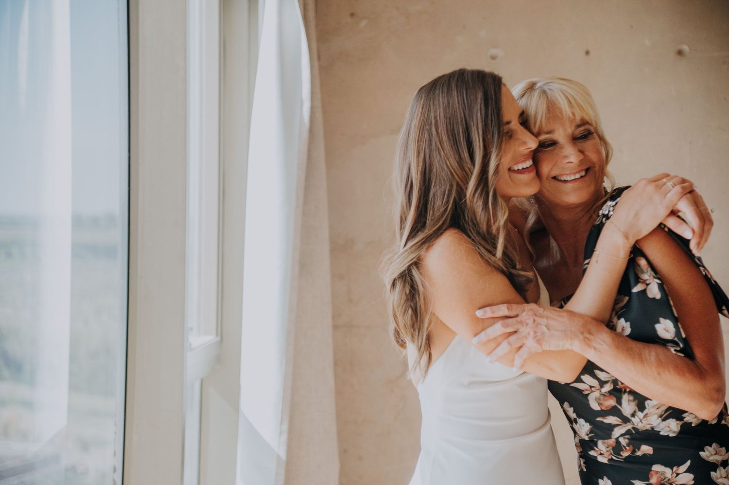Bride in white dress sharing a tender hug and kiss with older woman in floral dress near window.
