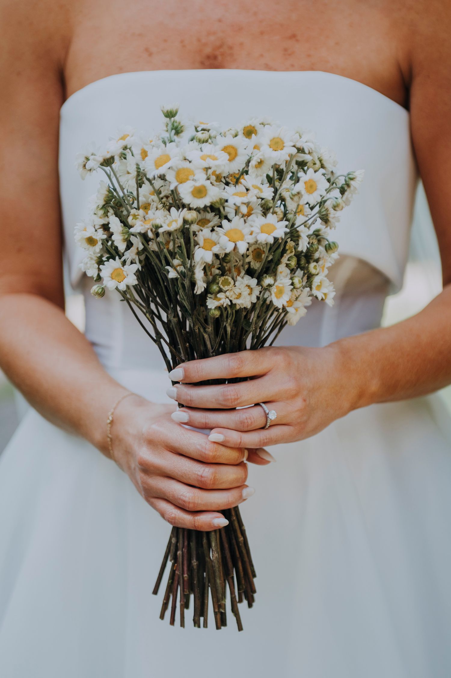 Bride in white strapless gown holding a rustic bouquet of small white daisies with dark stems, wearing a delicate ring.