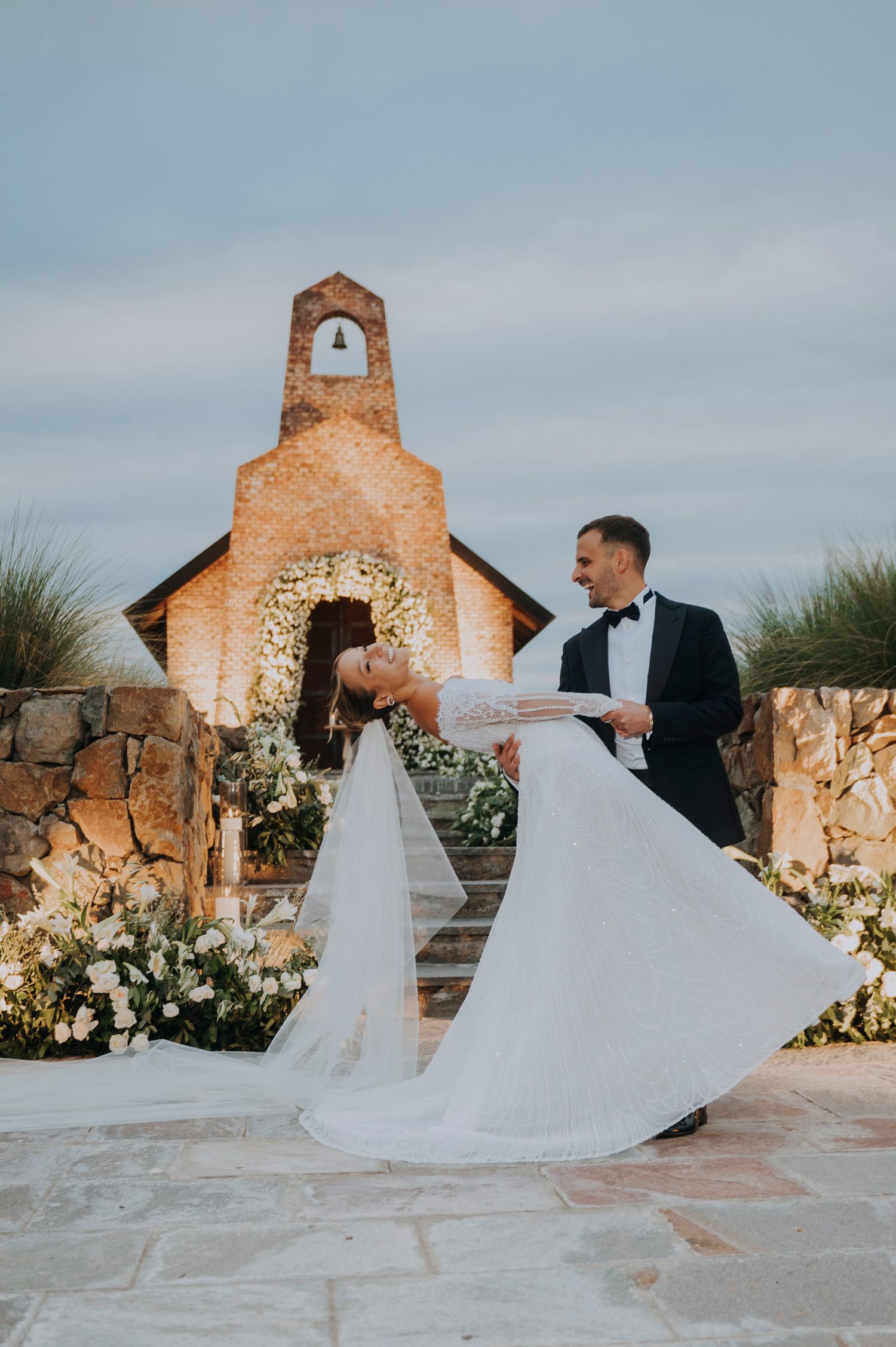 Bride and groom dancing outside illuminated stone chapel at dusk with white floral decorations.