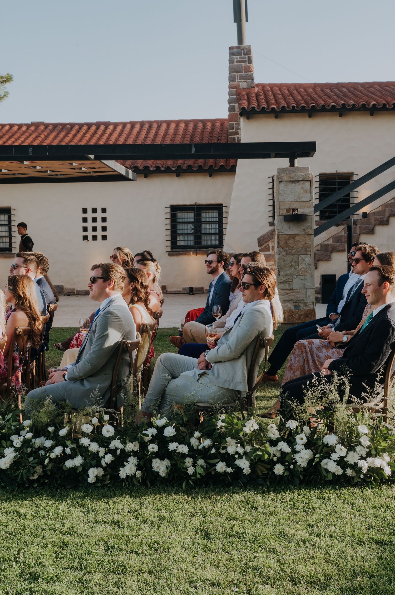 Wedding guests seated in chairs on green lawn watching outdoor ceremony near Spanish-style venue.