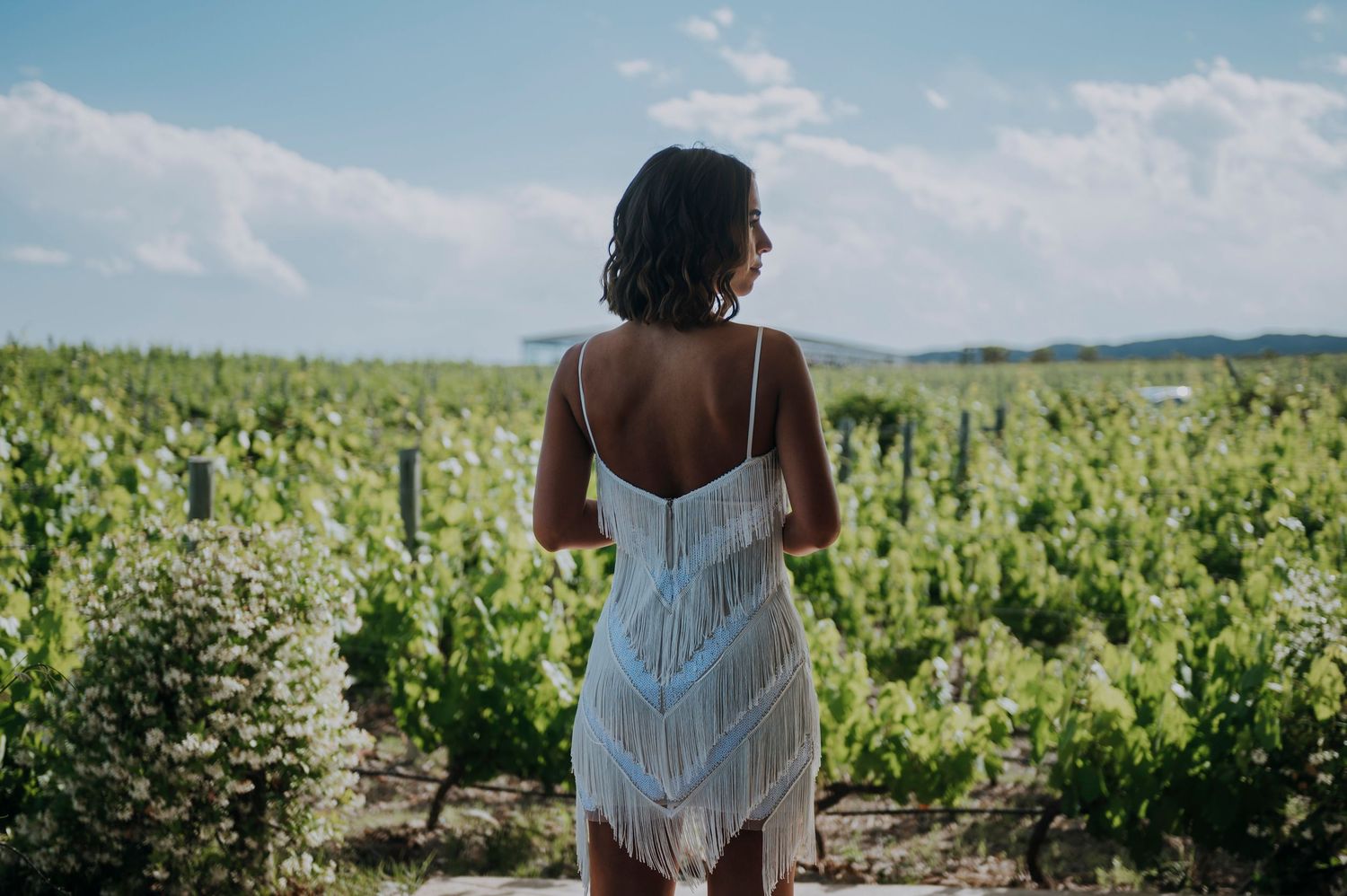 Woman in white fringed dress stands with her back to camera overlooking a lush vineyard on a sunny day.