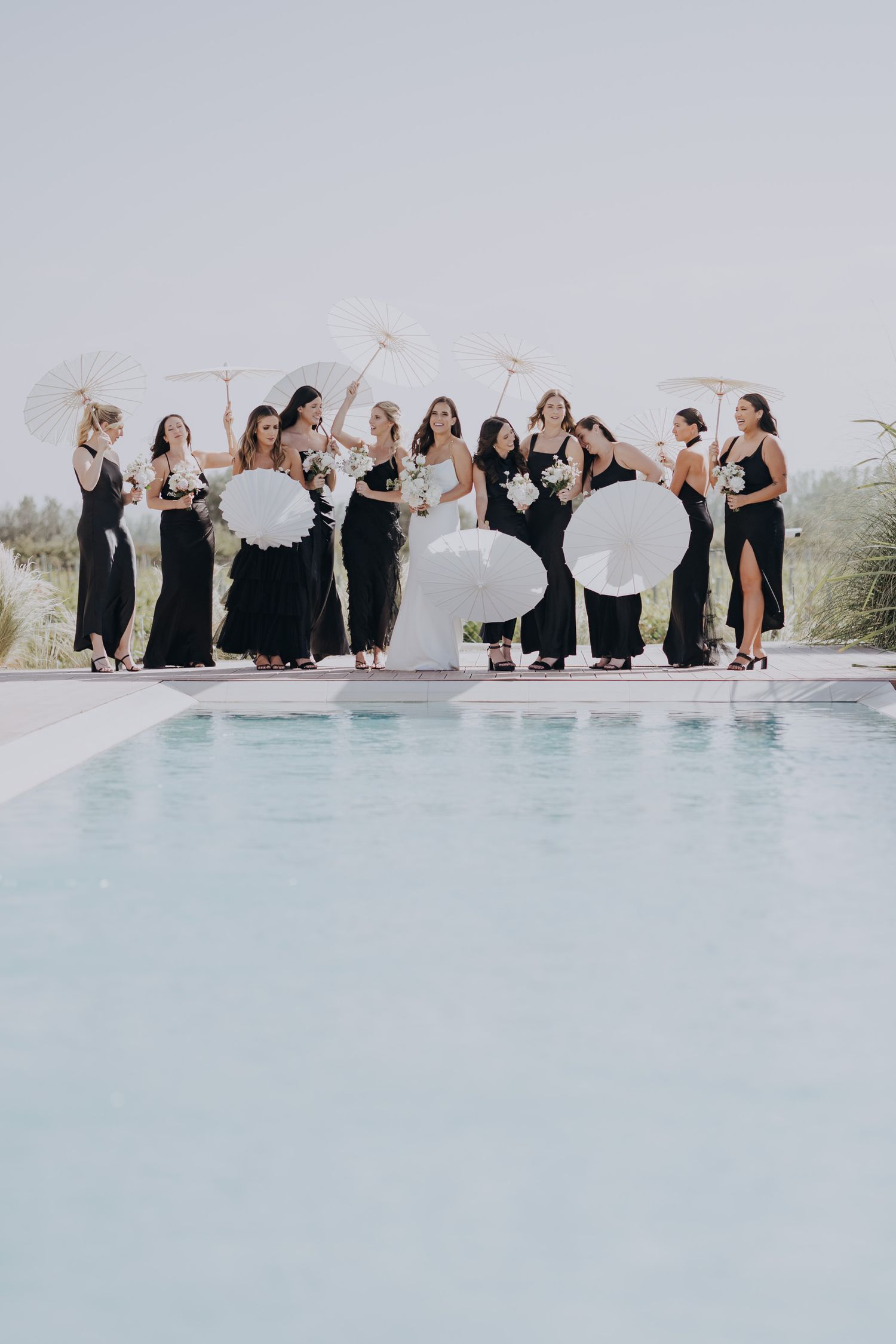Bridesmaids in black gowns holding white parasols and bouquets pose by a pool at an outdoor wedding.