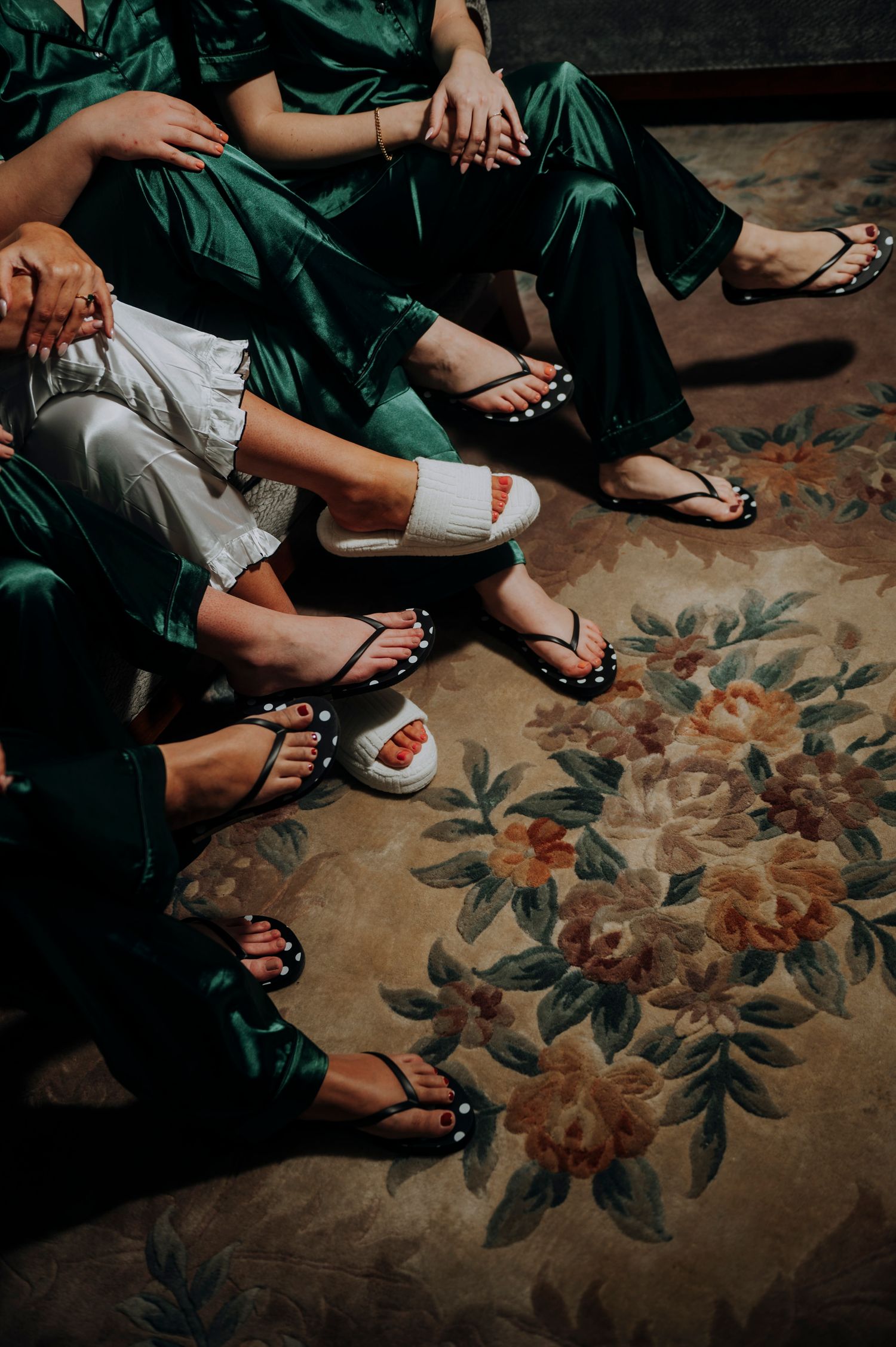 Bridesmaids in green satin robes sitting together wearing black flip flop sandals on floral rug.