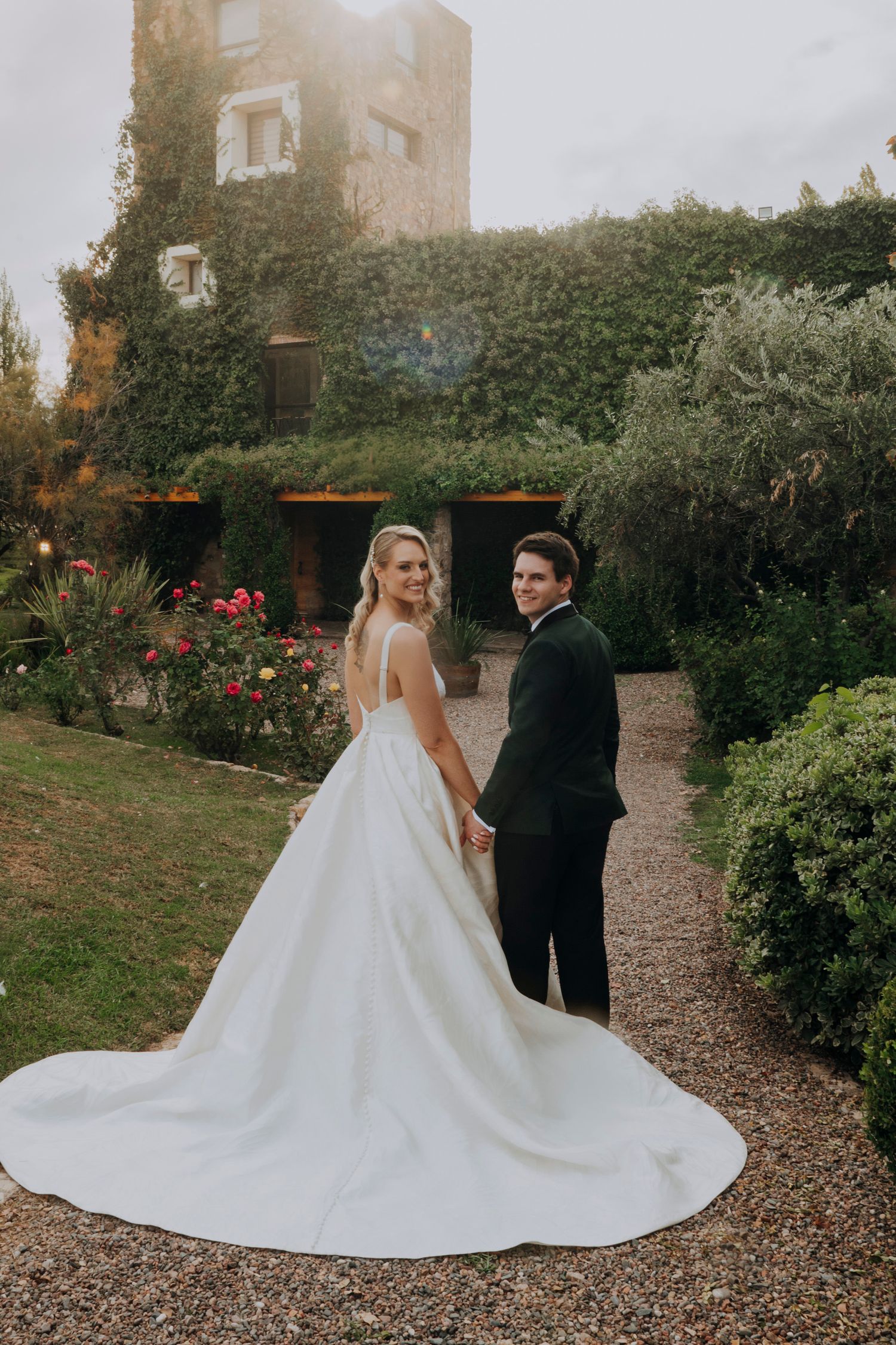 Bride and groom holding hands in lush garden, bride wearing dramatic cathedral train wedding gown.