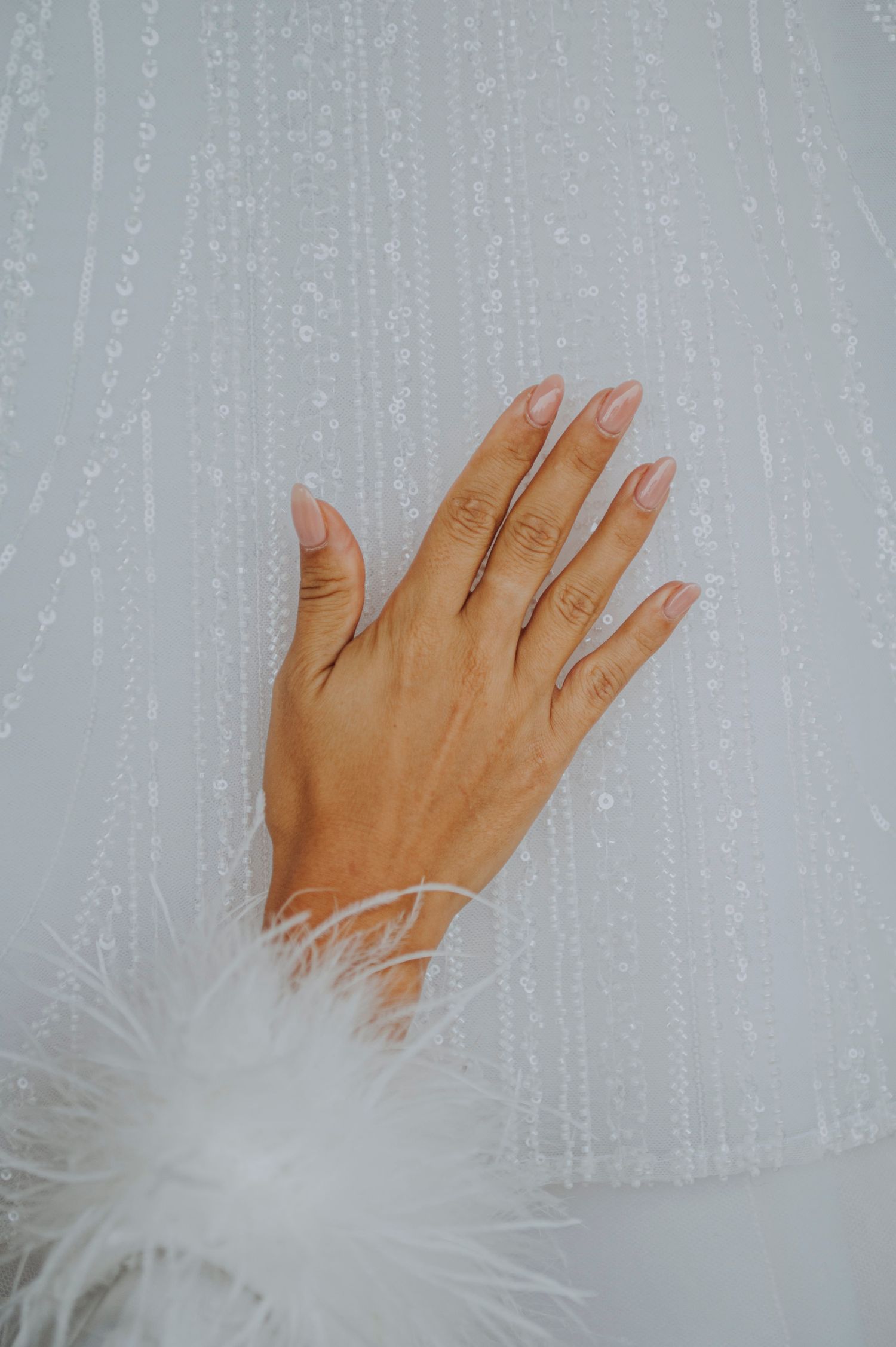 Close-up of a hand with pink almond nails resting on white beaded fabric with feather trim cuff.