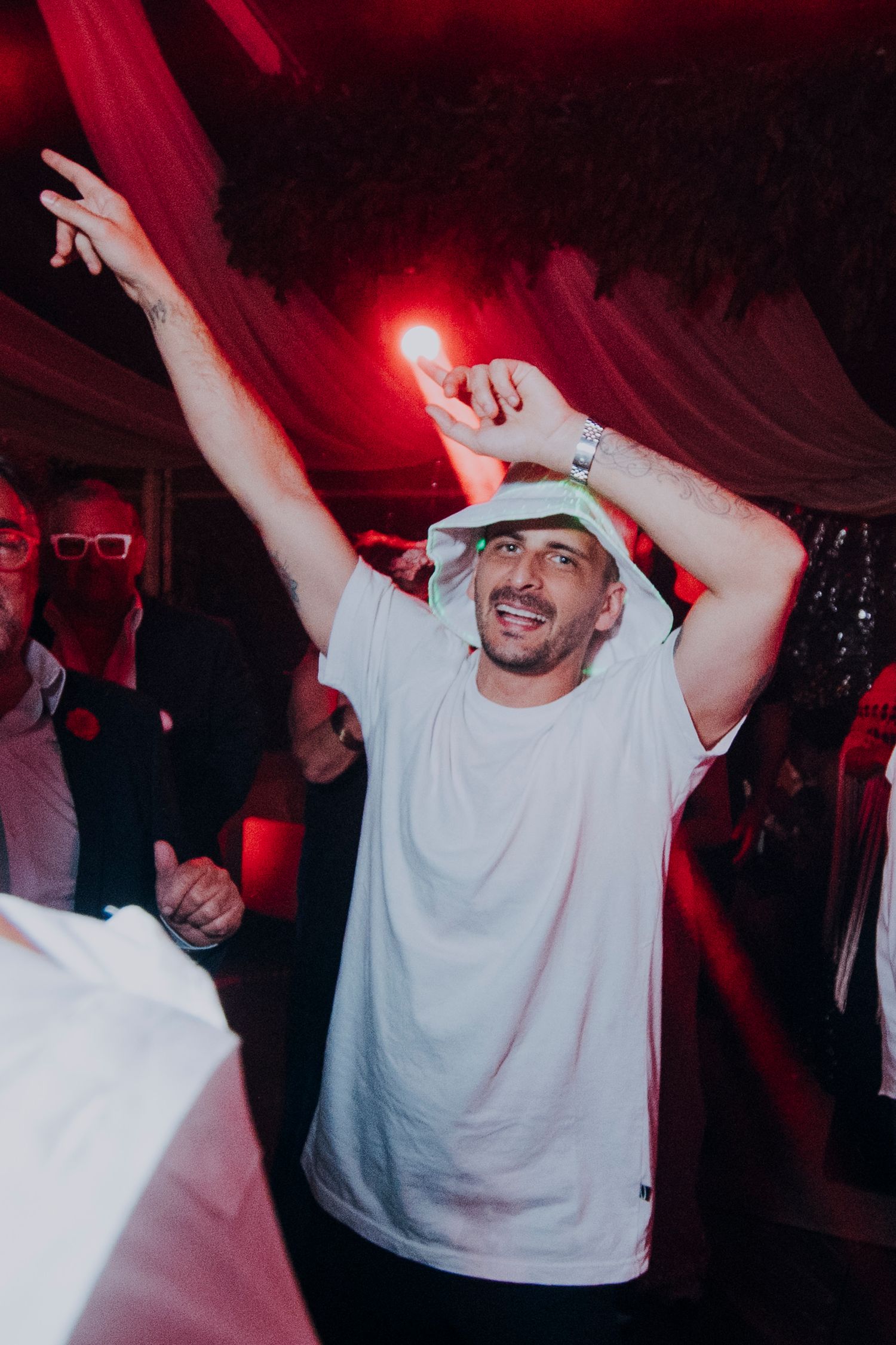 Guest in white t-shirt and bucket hat dances energetically at wedding party.
