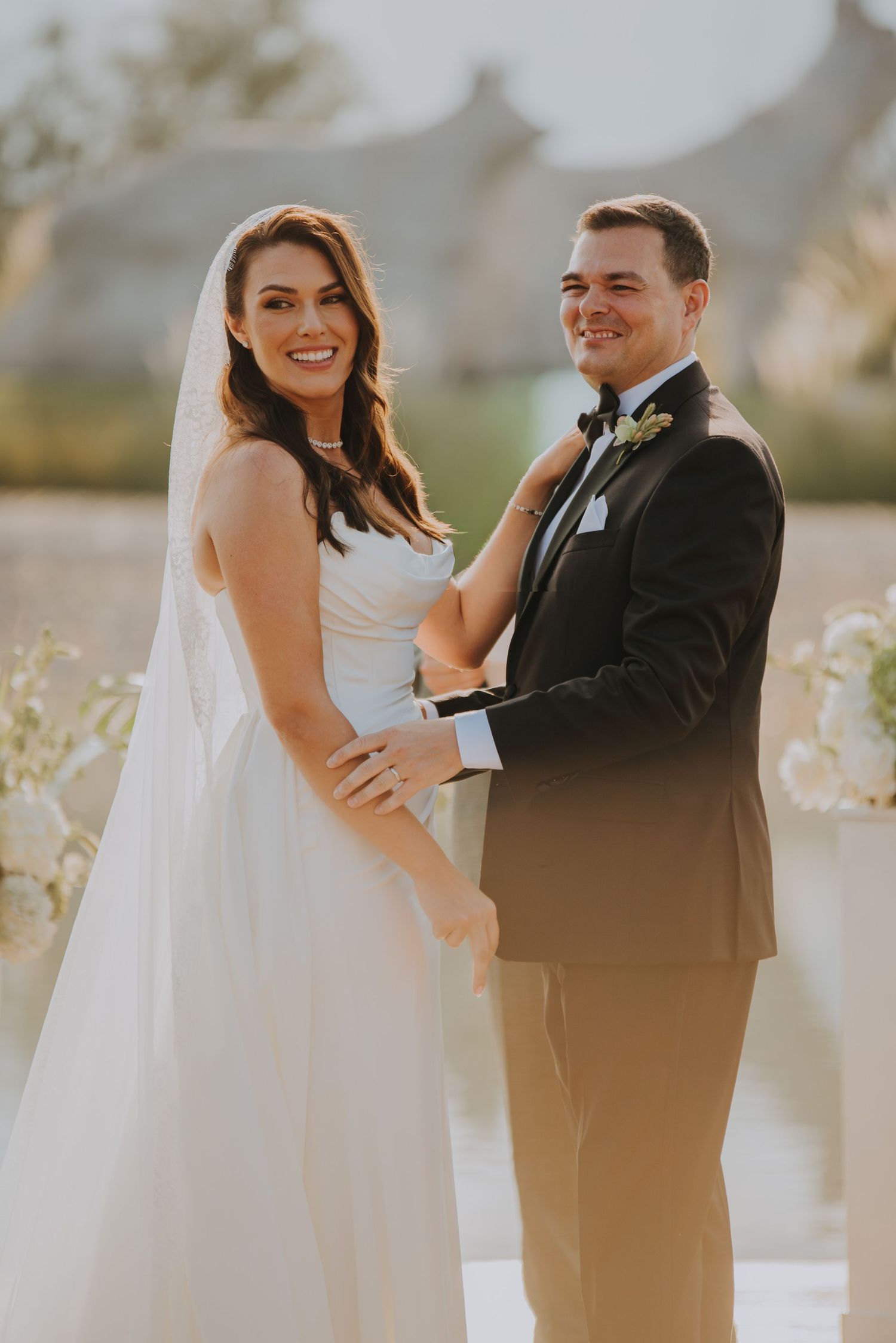 Smiling bride in white gown with veil and groom in black tuxedo posing together at outdoor wedding ceremony.