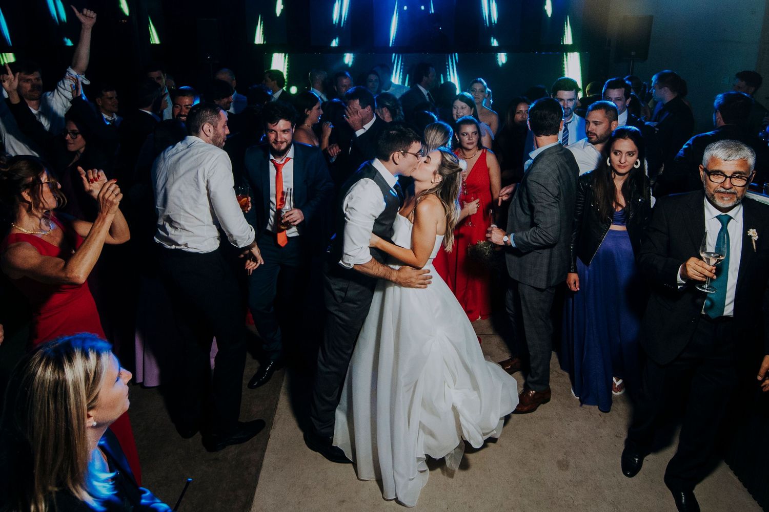 Bride and groom share passionate kiss while guests dance around them at wedding reception.