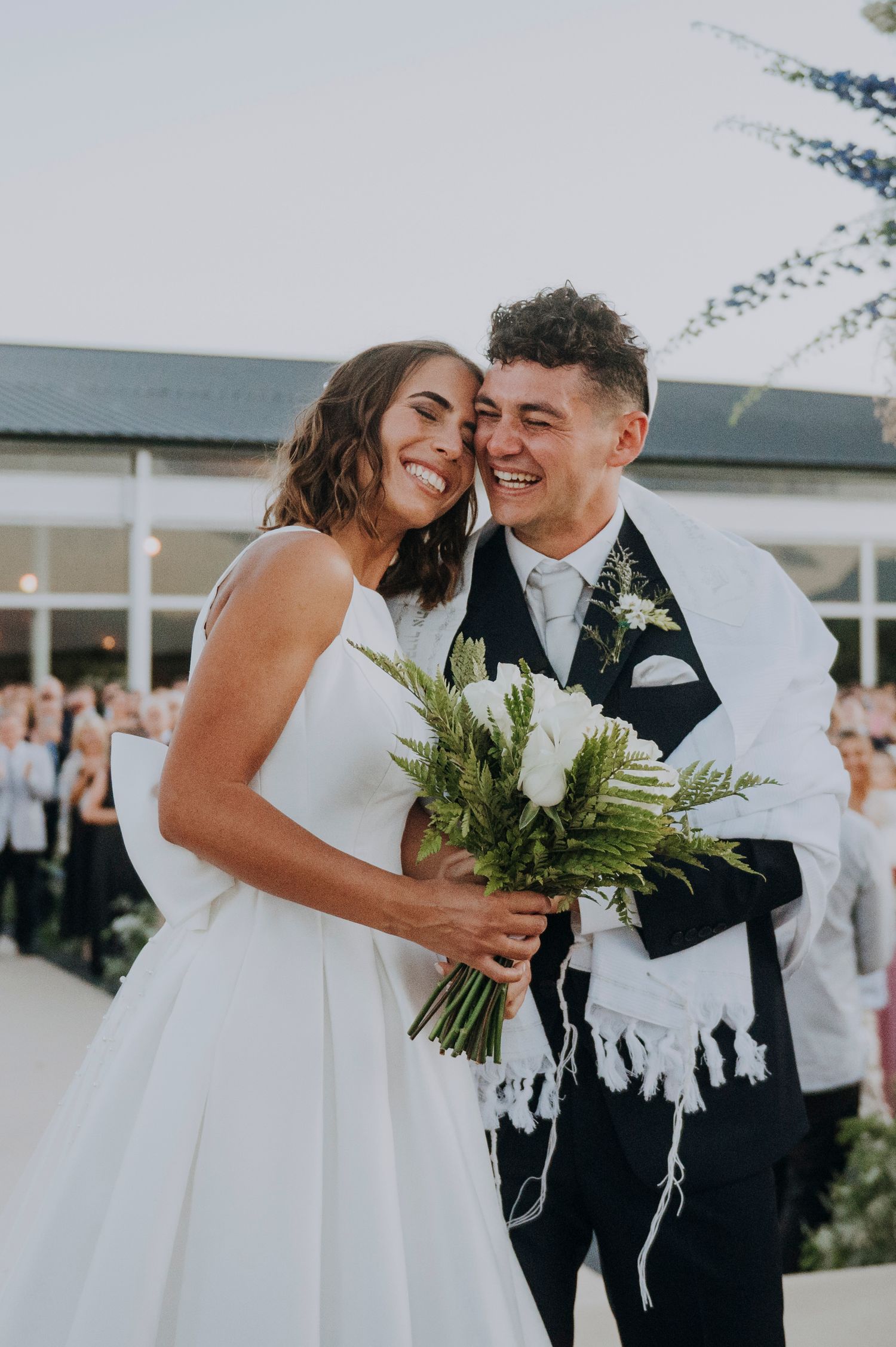 Happy bride and groom embrace after Jewish wedding ceremony in Mendoza holding white bouquet under tallit.