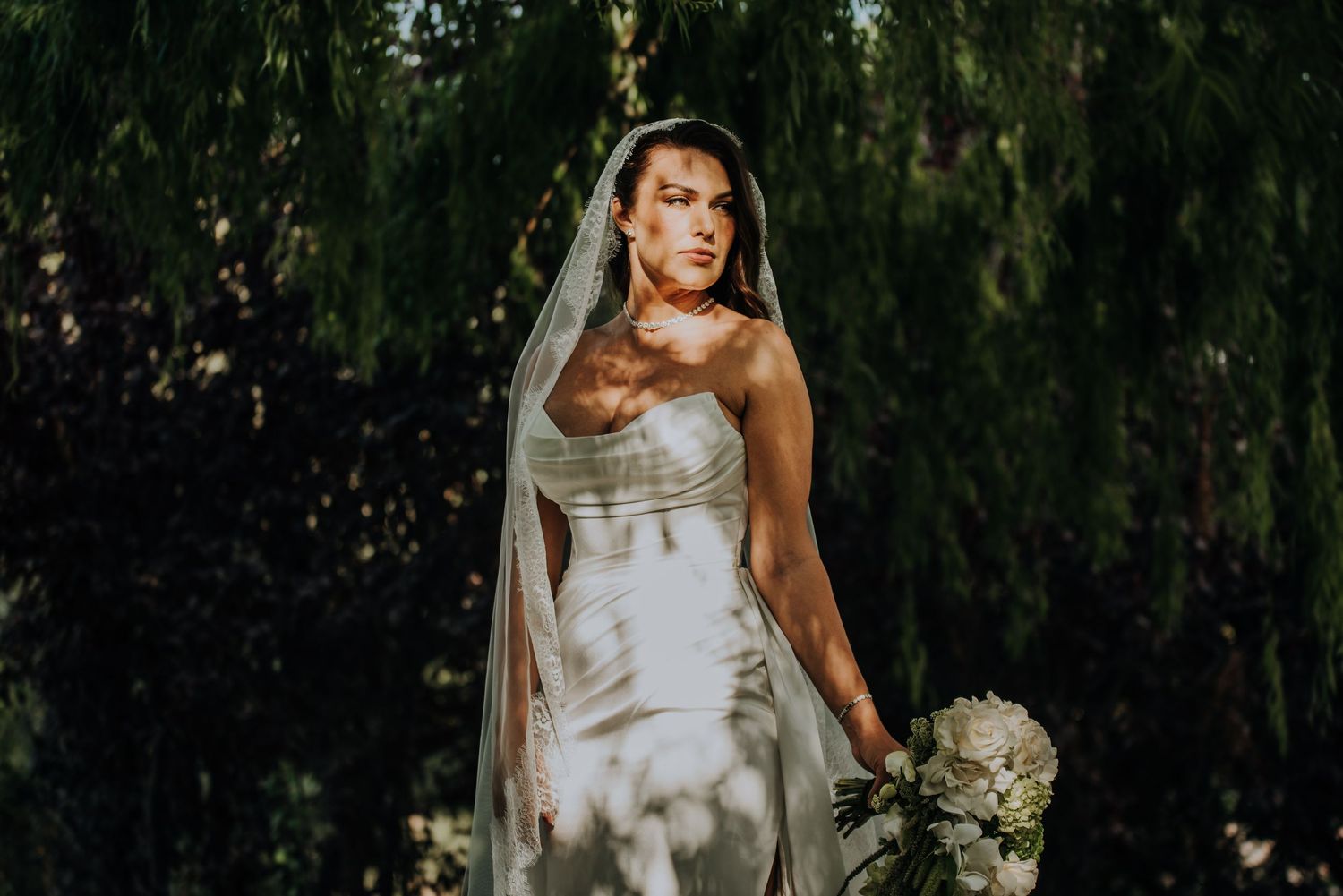 Bride in strapless white gown and veil holds bouquet outdoors under weeping willow tree in dappled sunlight.