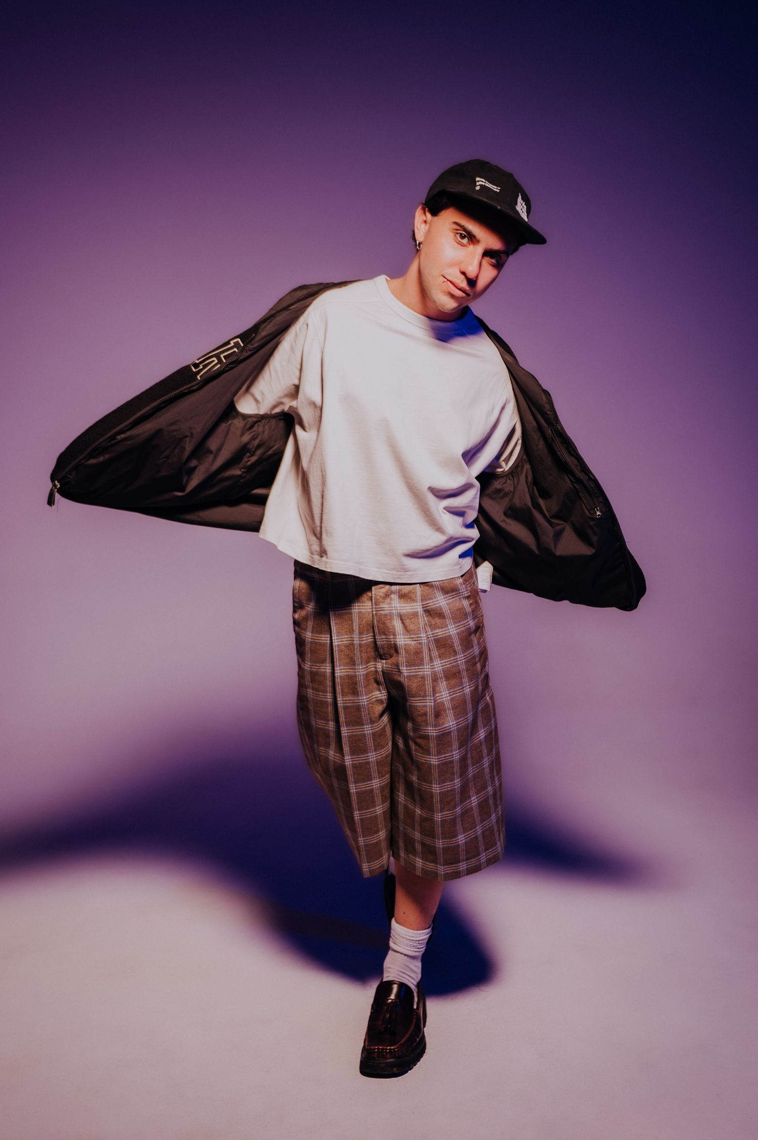 Igna Farres Young man in cap, white sweatshirt, and plaid shorts posing with jacket against purple background.
