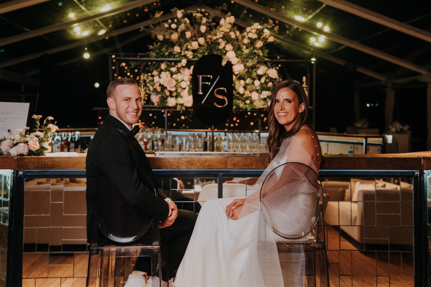 Bride and groom smiling at a decorated reception bar with string lights and monogram signage.