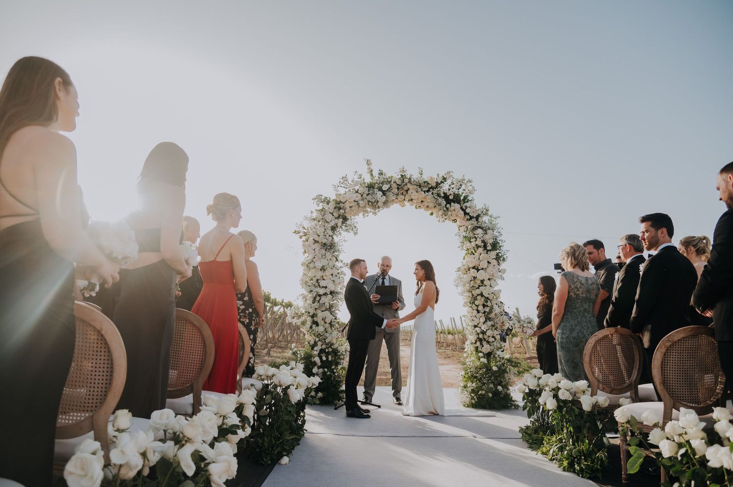 Couple exchanges vows under a floral arch surrounded by guests at an elegant outdoor wedding ceremony.