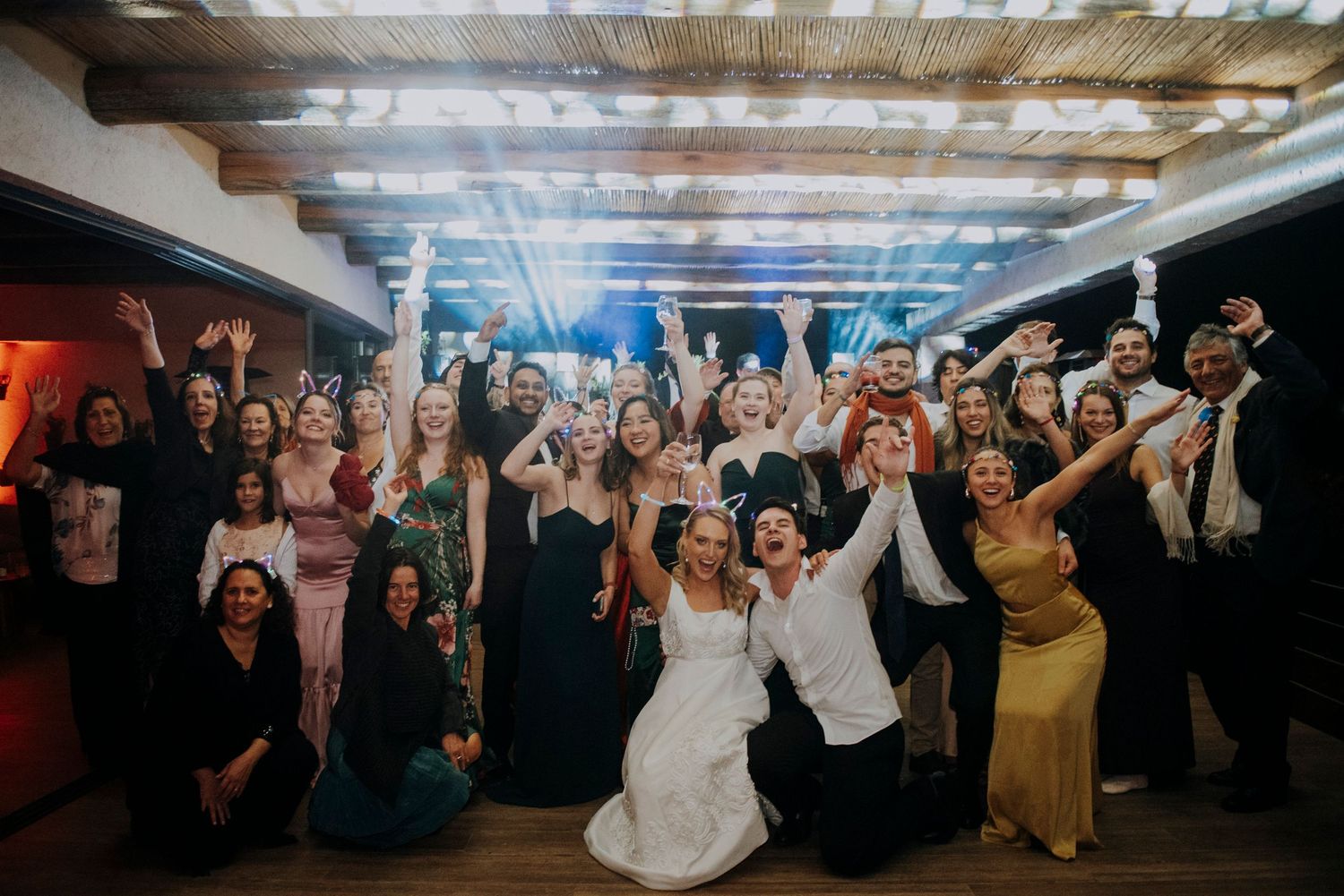 Bride and groom celebrating joyfully with large group of wedding guests on illuminated dance floor.