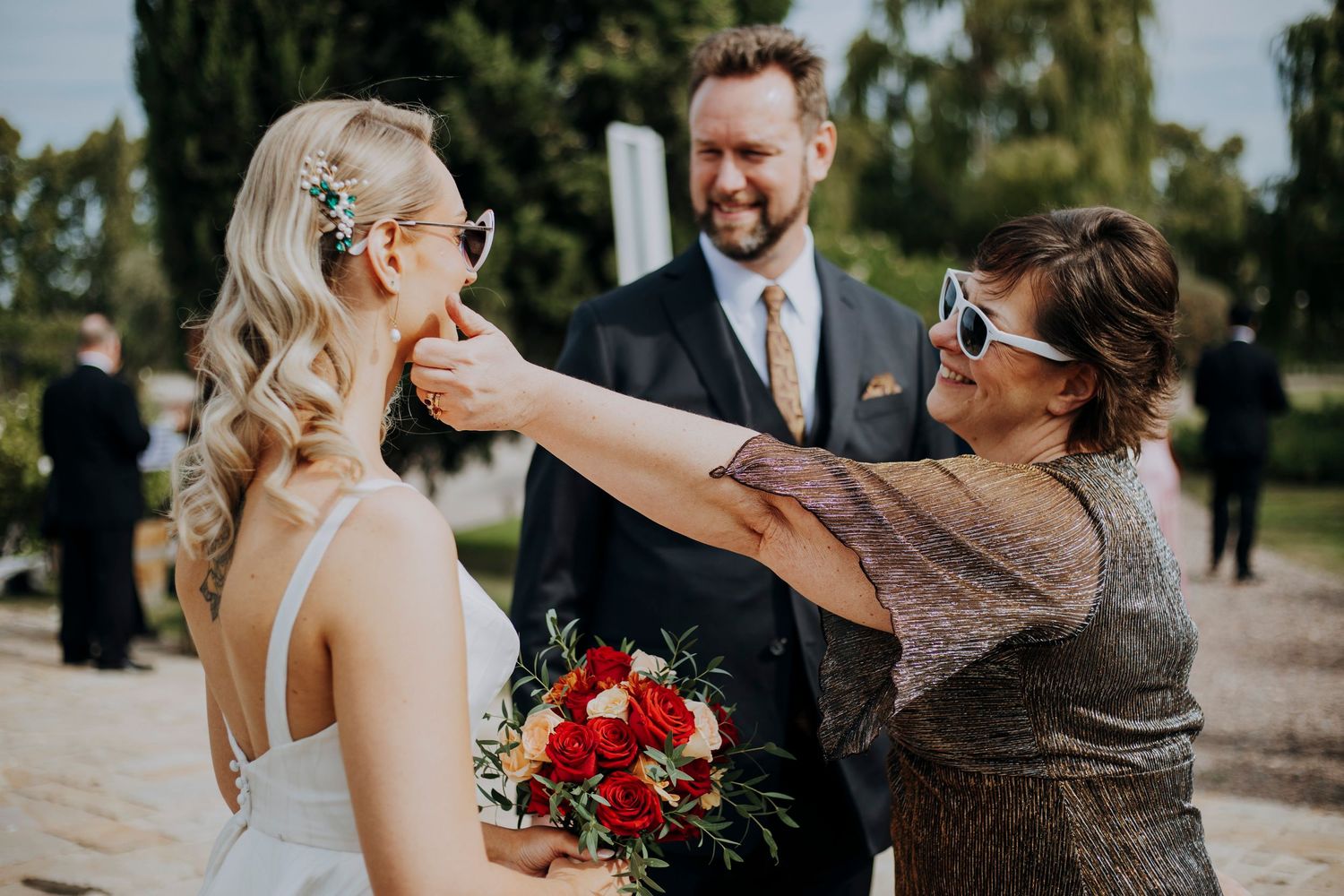 Woman placing heart-shaped sunglasses on blonde bride holding red bouquet at outdoor wedding.