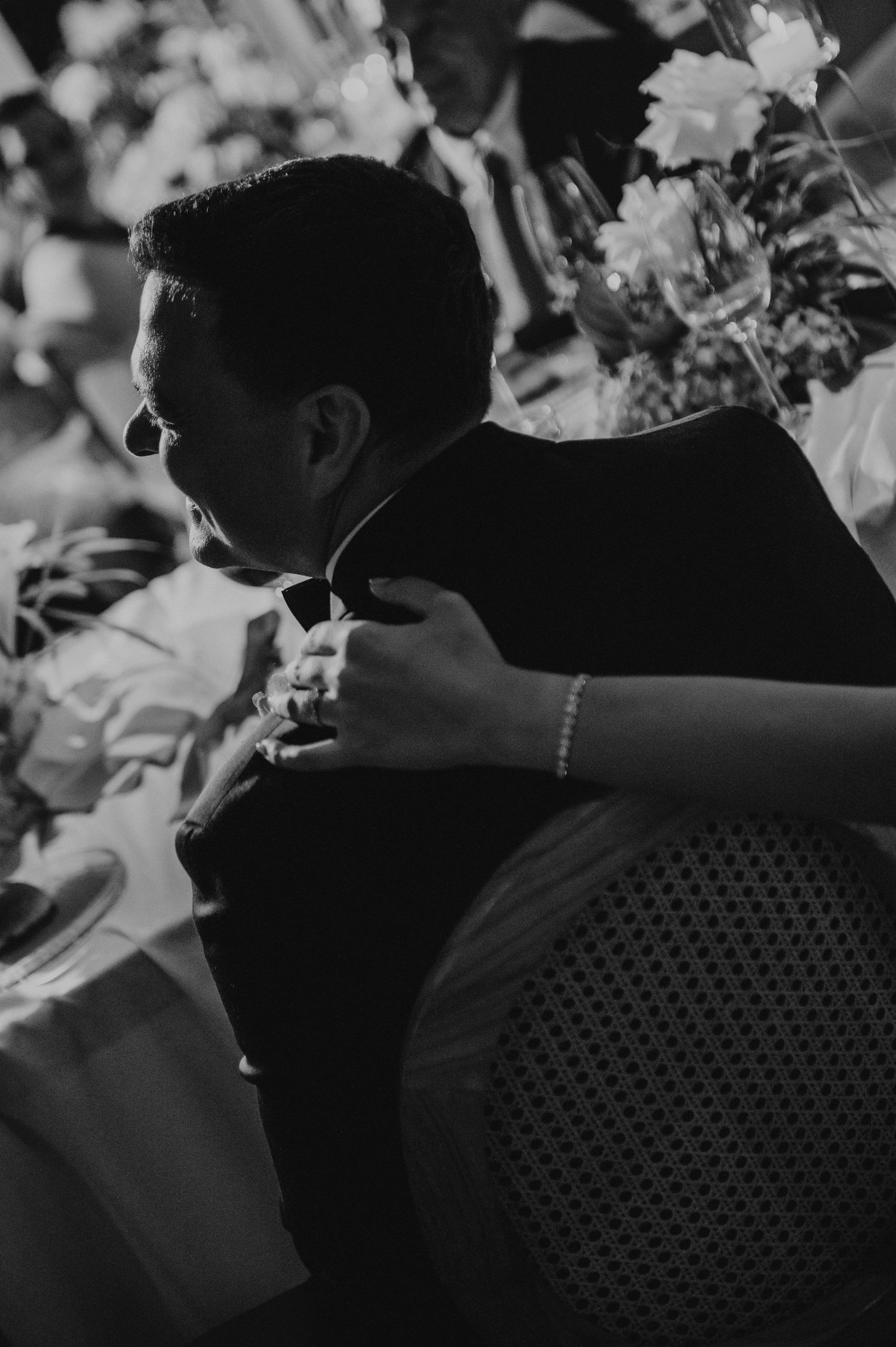 Black and white photo of a groom in a tuxedo being embraced at a wedding reception with floral arrangements.