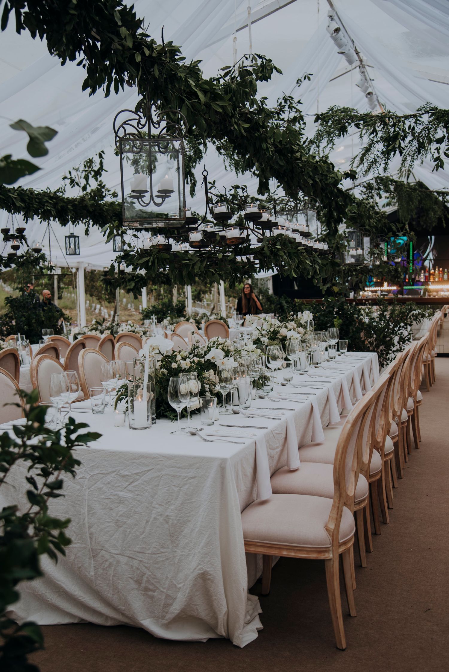 Elegant wedding reception under tent with long white tables, greenery ceiling, and lanterns.