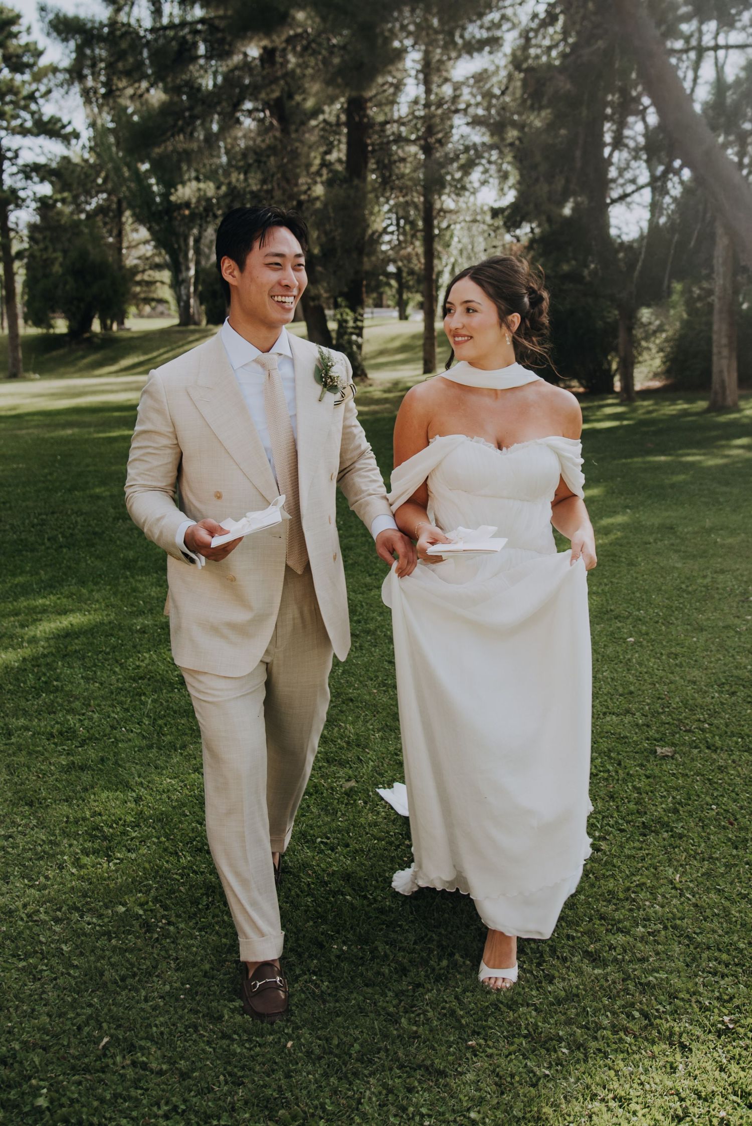 Smiling newlyweds walk hand-in-hand on green lawn, groom in beige suit, bride in off-shoulder white gown.