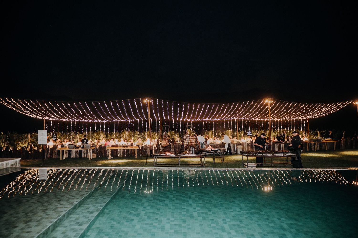 Stunning outdoor wedding reception at night with string lights over pool reflecting warm golden glow.