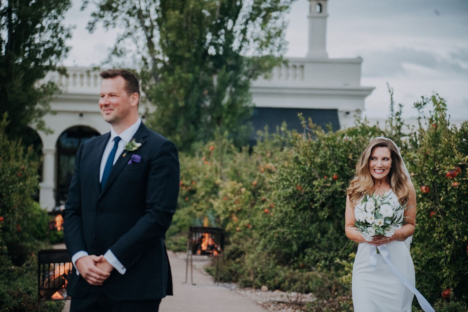 Bride in white gown holding bouquet smiles at groom in navy suit during outdoor wedding ceremony.