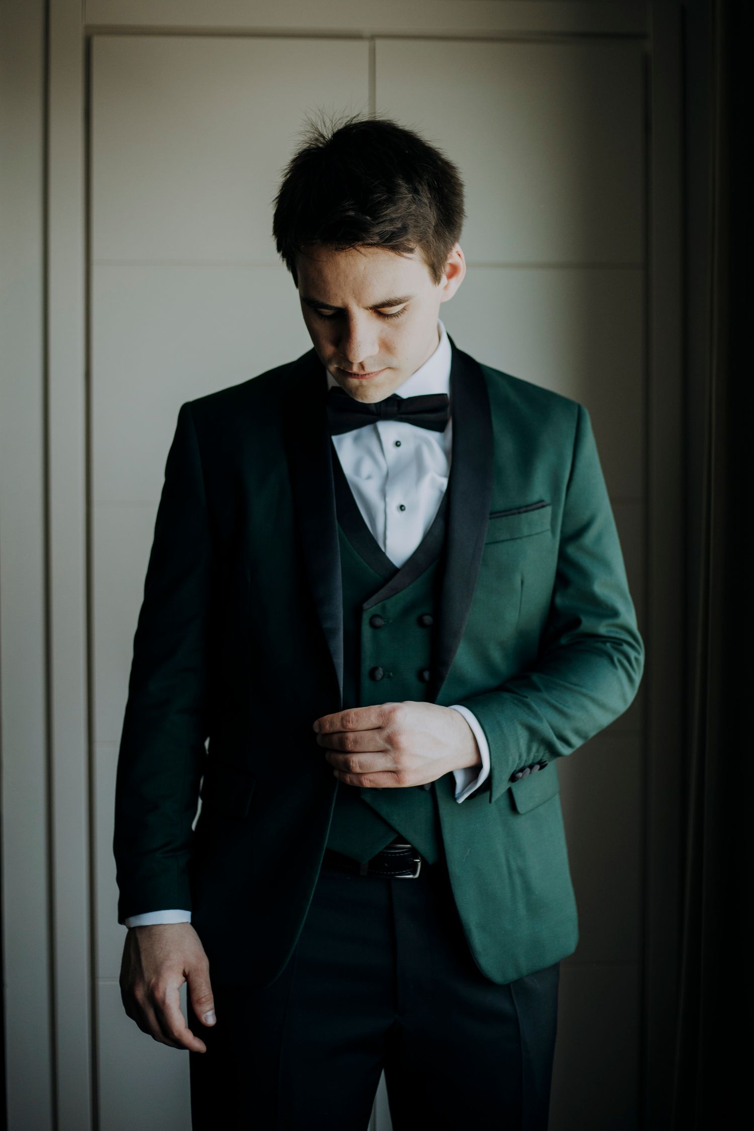 Groom in a dark green tuxedo with black bow tie adjusting his jacket button before the wedding ceremony.