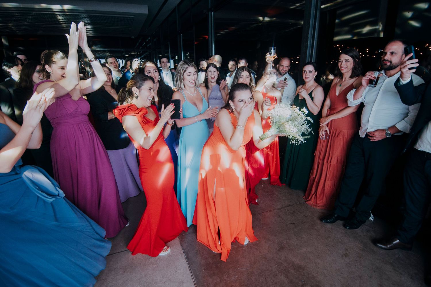 Excited bridesmaids in colorful gowns catching bouquet at indoor wedding reception celebration.