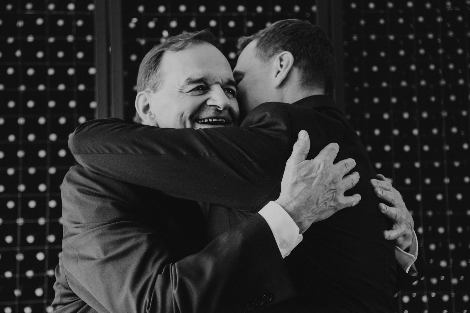 Two men in suits share a warm embrace and laugh together at a wedding reception.