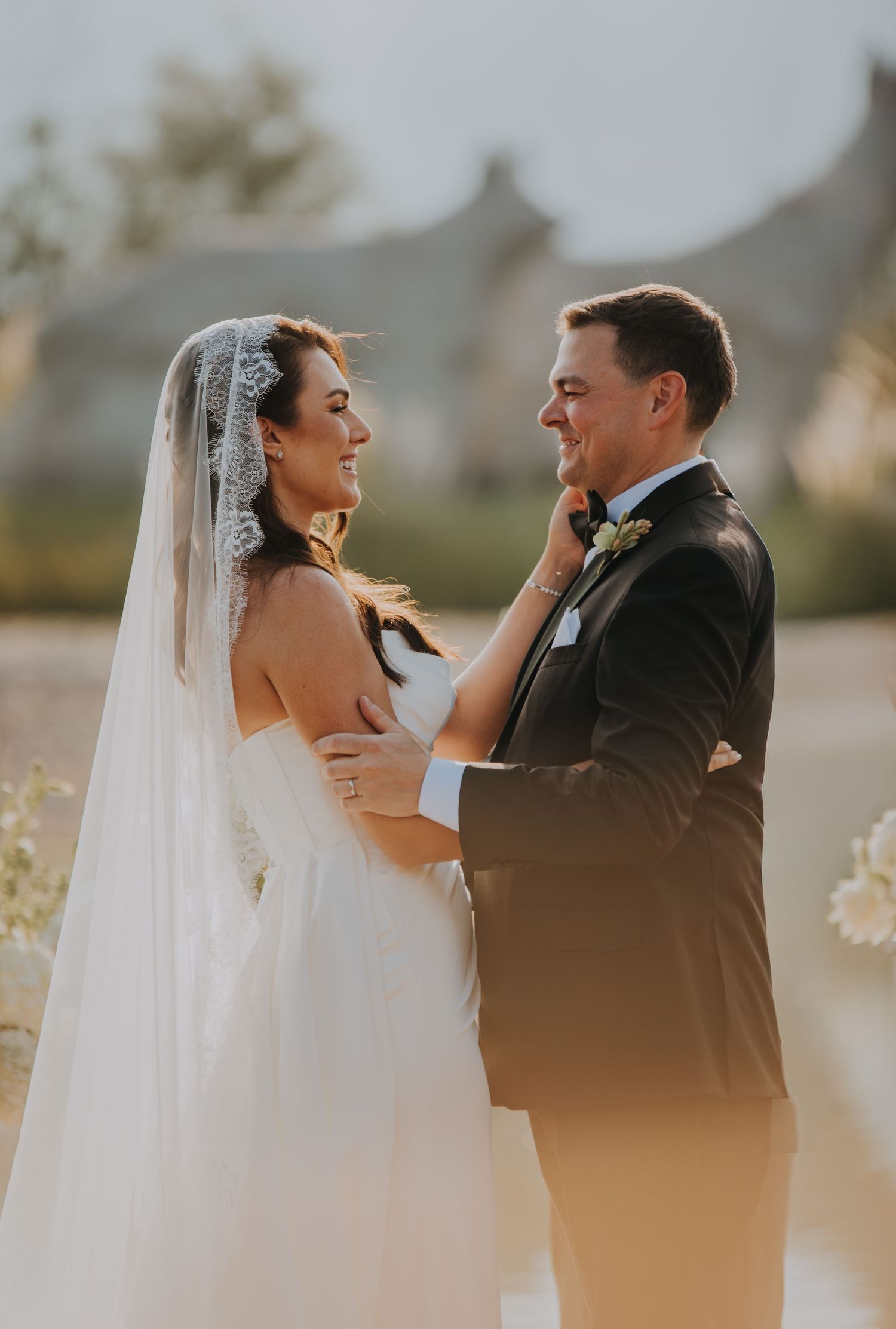 Smiling bride in white gown and veil gazes at groom in black tuxedo during golden hour outdoor wedding.