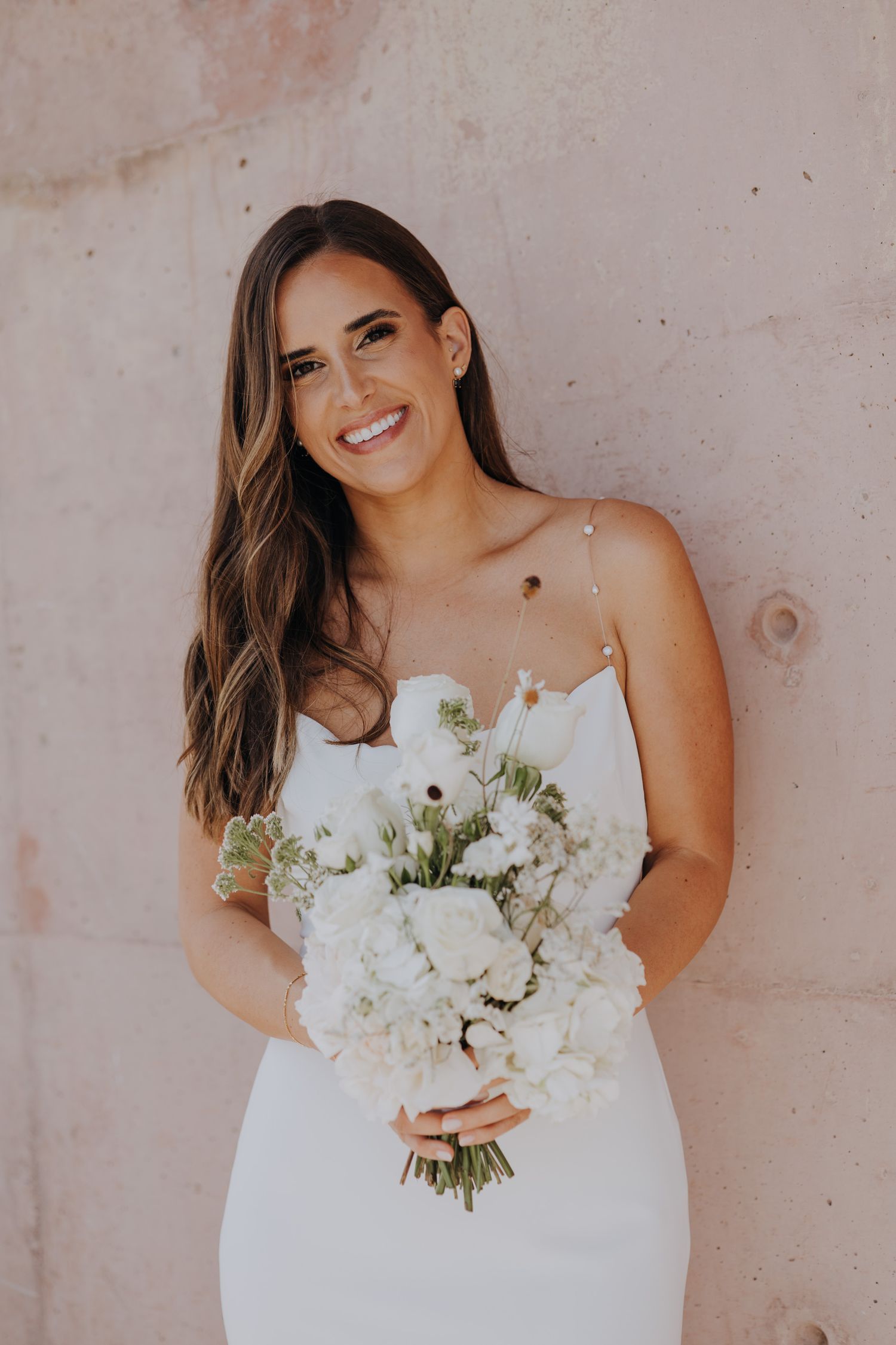 Radiant bride in white dress holding white bouquet smiling against a blush stone wall.