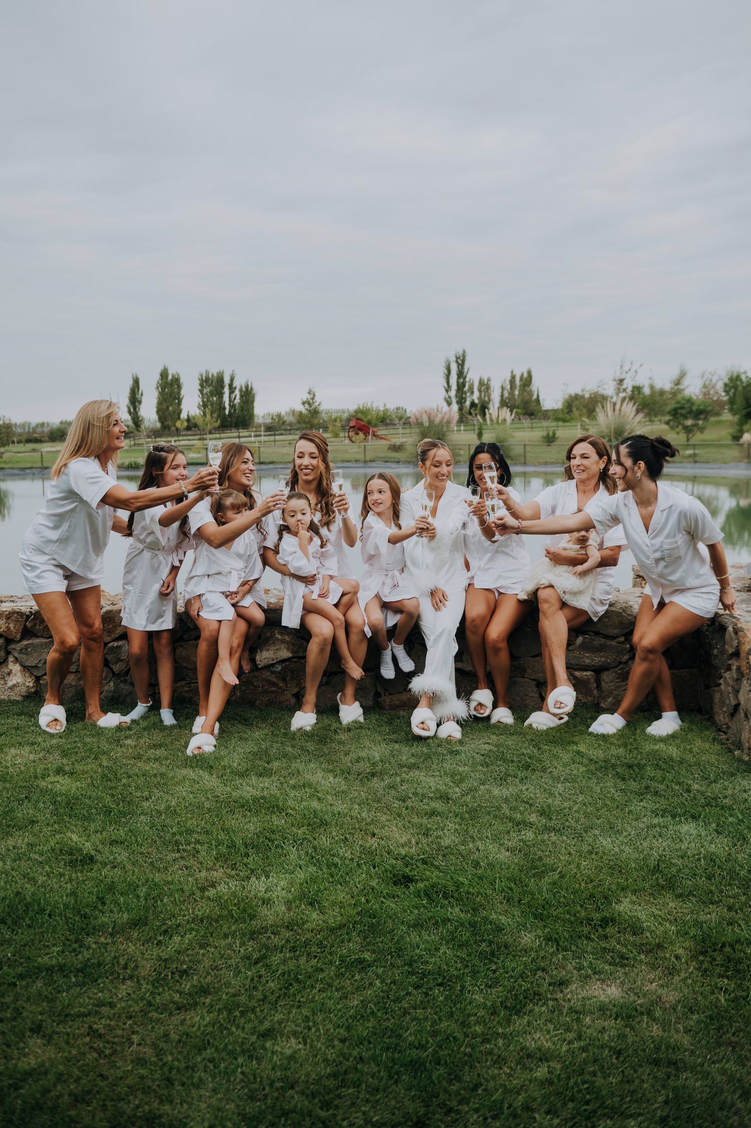 Bridesmaids in matching white robes celebrate with the bride outdoors by a pond on a wedding day.
