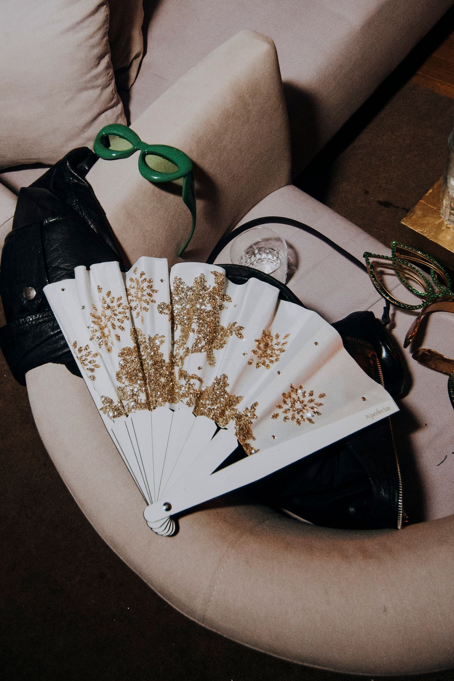 Decorative white and gold glitter hand fan displayed on a chair with green sunglasses and purse.