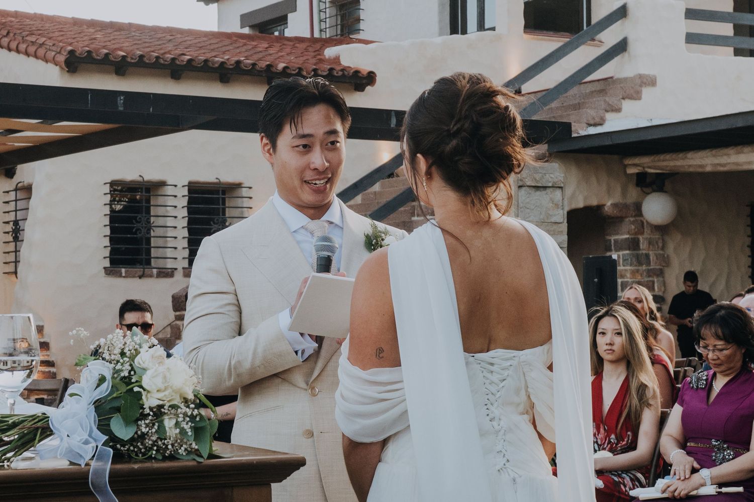 Groom in beige suit reads vows to bride at outdoor wedding ceremony with guests.
