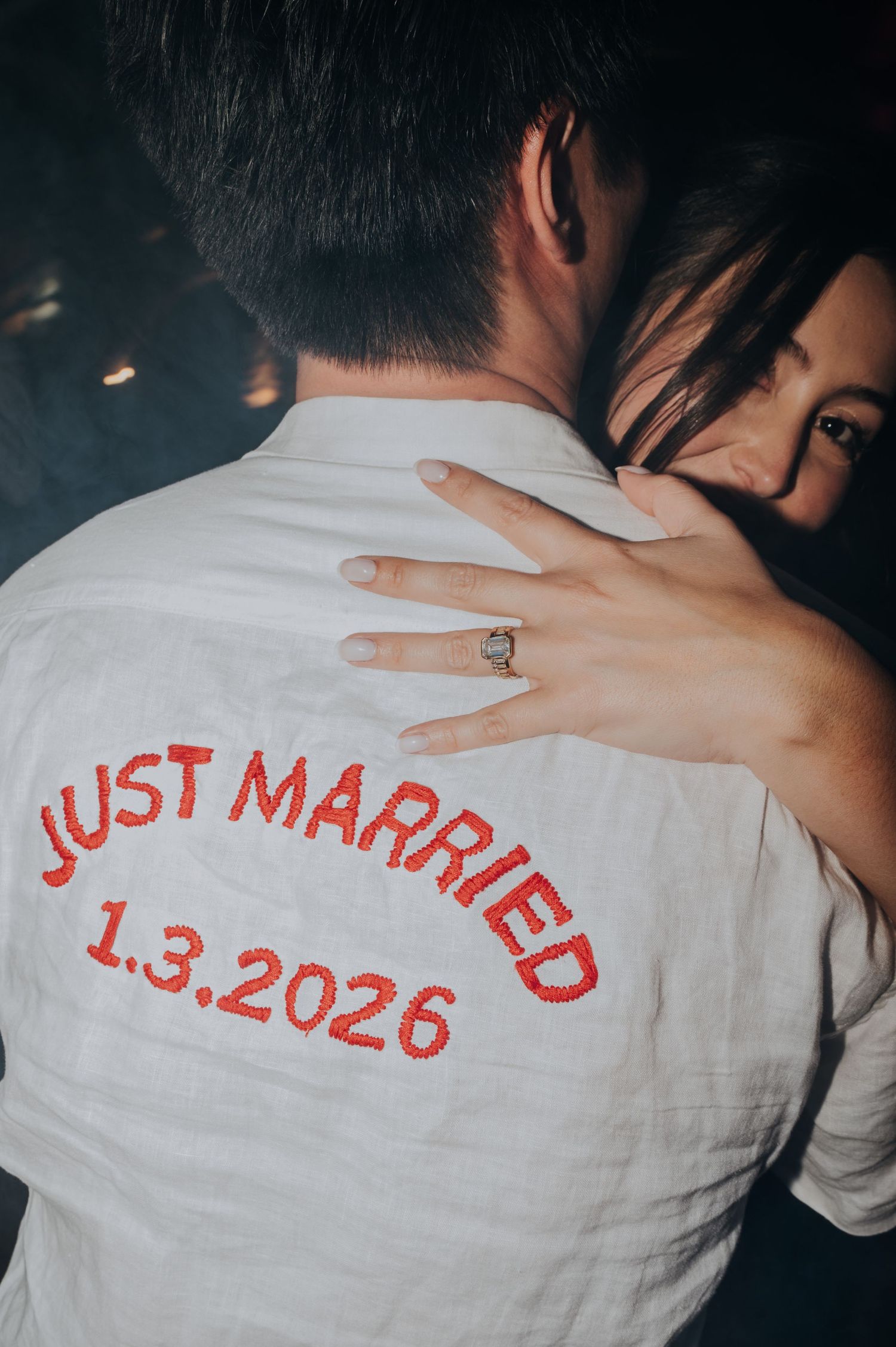 Newlywed groom wearing a white Just Married 1.3.2026 embroidered shirt while embracing his bride.