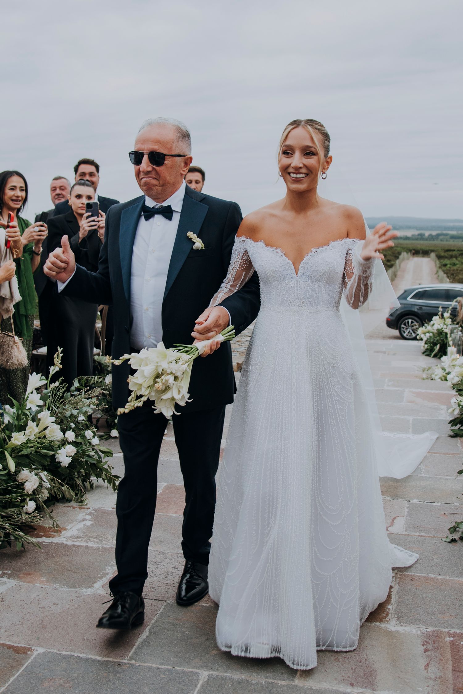 Bride in off-shoulder lace wedding gown walks down aisle with older man in tuxedo.