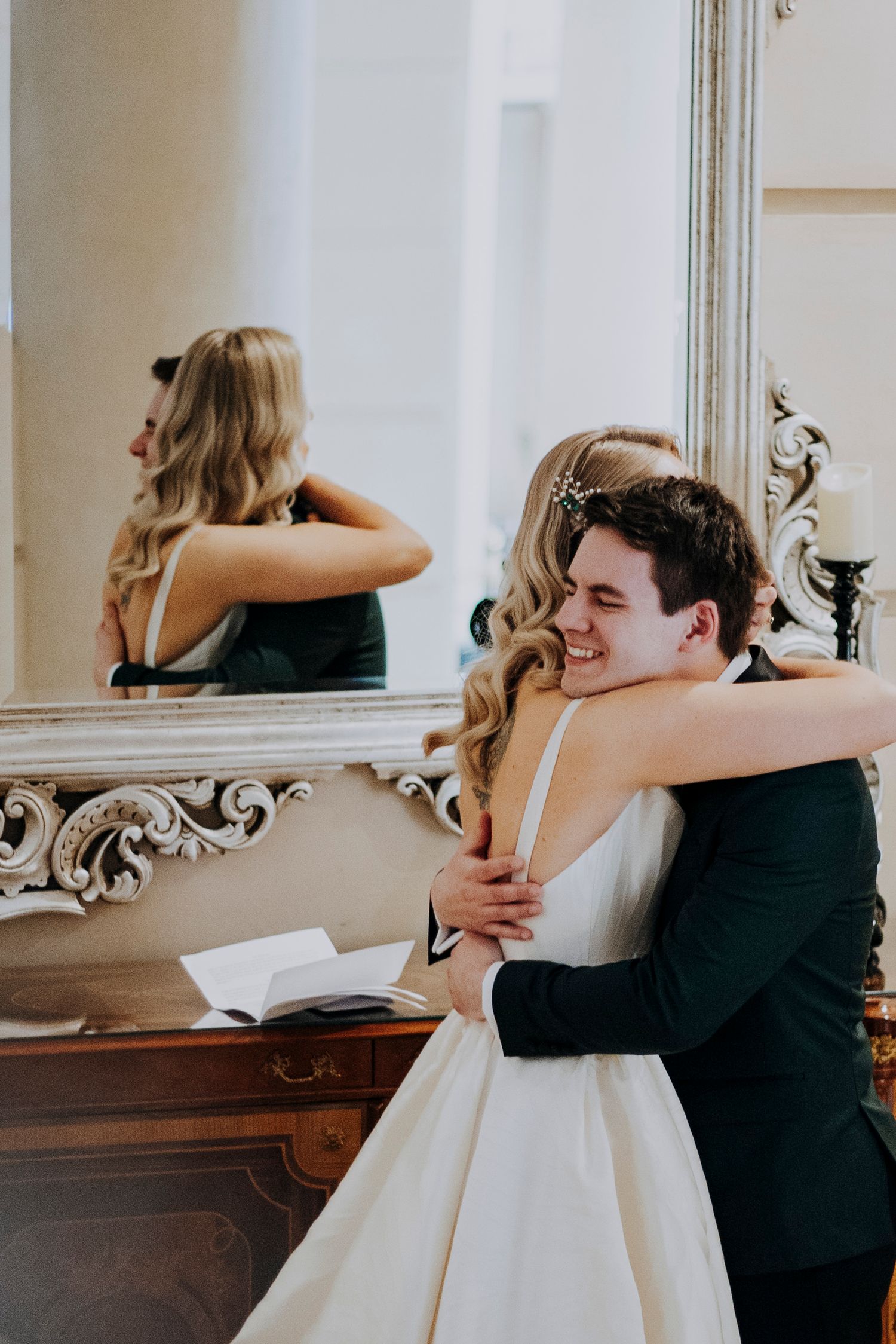 Smiling groom in green tuxedo embraces bride in white gown near ornate mirror at wedding.