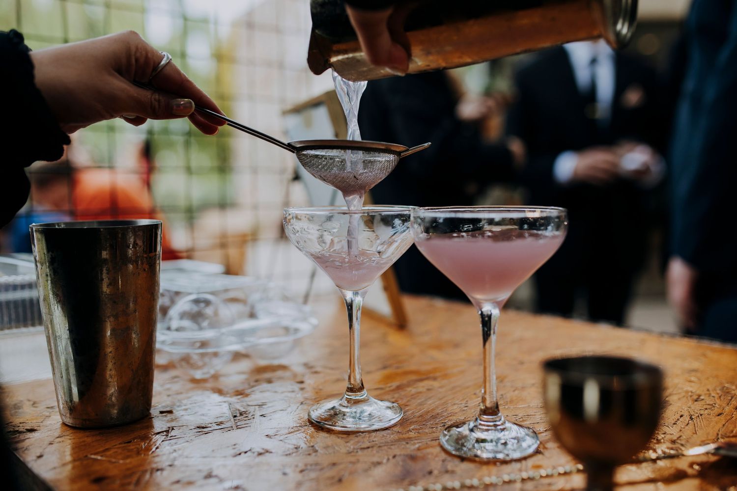 Bartender straining pink cocktails into elegant coupe glasses on rustic wooden bar.