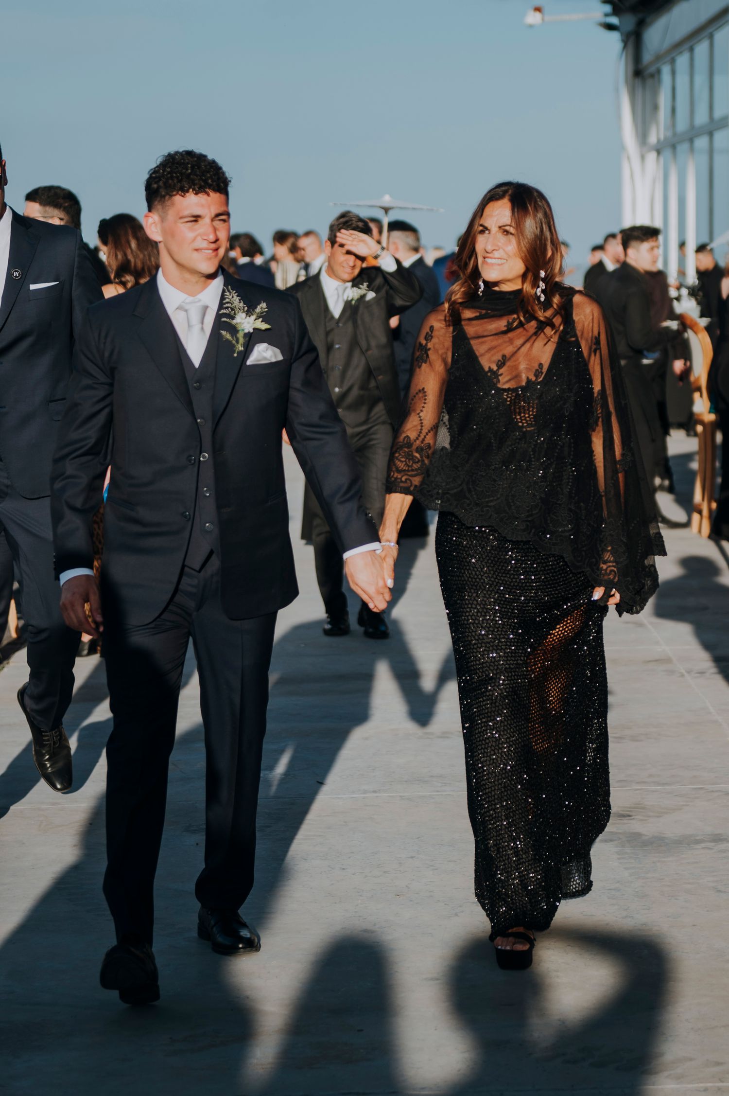 Elegant couple walks hand in hand outdoors, groom in black suit, bride in black lace gown.